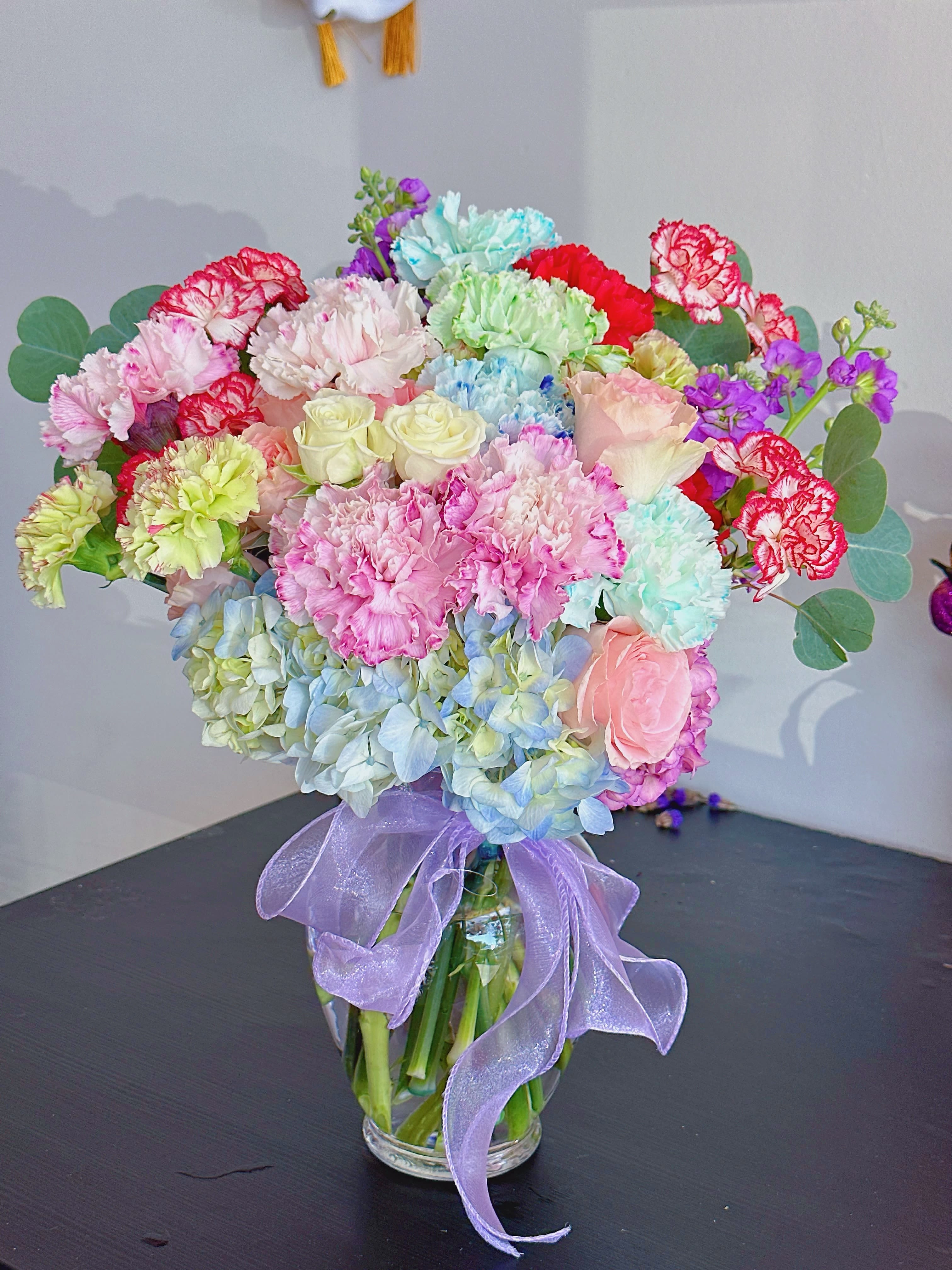 Colorful bouquet of carnations, roses, and hydrangeas in a glass vase with a purple ribbon