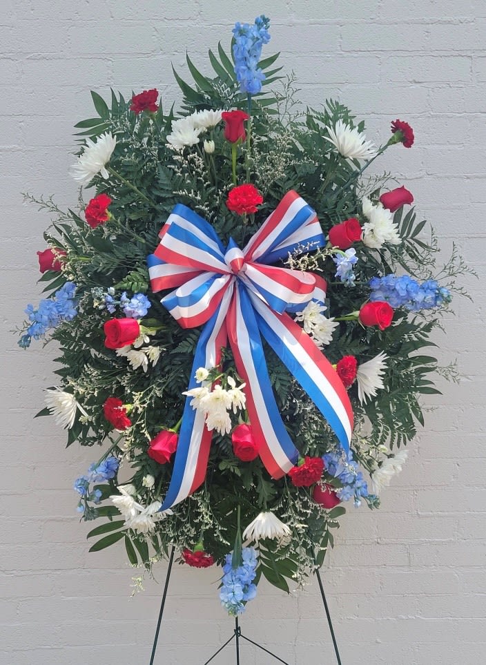 Red, White and Blue Standing Spray  - A full circular standing spray that reads as both patriotic and reverent, this red, white and blue standing spray is designed for sympathy and funeral display. The dense, symmetrical wreath sits on a floral easel, with a large red, white, and blue ribbon bow at the center that lends a ceremonial, honoring presence-perfect for commemorating a veteran, public servant, or any loved one with national ties.  The arrangement combines red roses and red carnations with white daisy-like blooms, blue delphinium and accents of green foliage for contrast. Flowers are arranged to extend outward evenly for a formal, full silhouette; trailing delphinium adds vertical movement while the ribbon anchors the composition. Supplied on a sturdy floral easel with a red, white and blue ribbon, this spray arrives ready for immediate display.  Appropriate for memorial services, graveside ceremonies, or public commemorations, this piece communicates respect and support for the person you're honoring. 