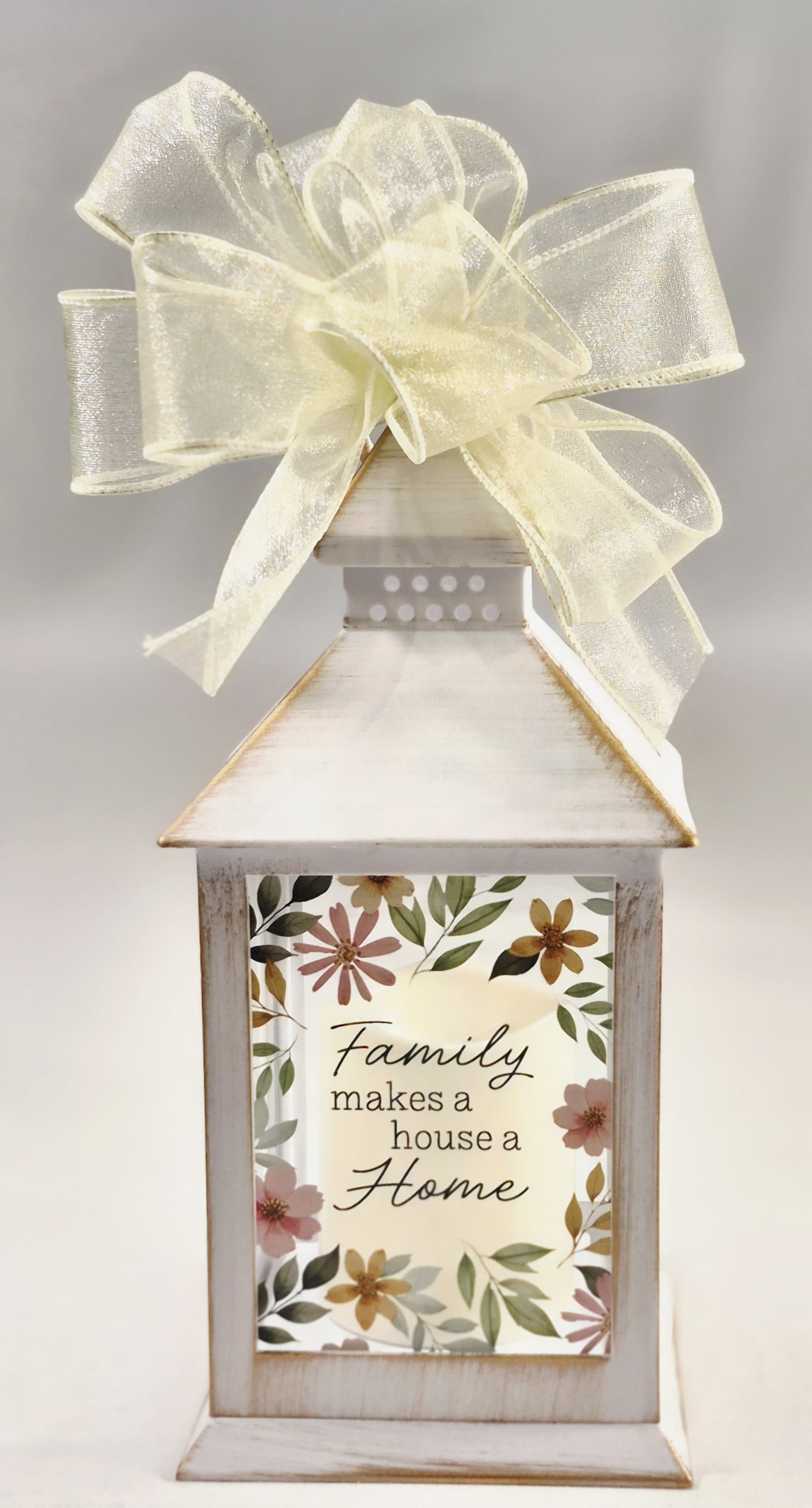 Family Makes a House a Home Lighted Lantern - A distressed white wooden lantern topped with a sheer yellow bow this lighted lantern is a charming home décor gift that carries the message "Family makes a house a Home" right on its front panel. Designed as a wedding or housewarming gift, the sentimental plaque and soft neutral palette-with delicate pink and green floral accents-make it an inviting touch for entryways, mantels, or bedside tables.  The lantern features a farmhouse-inspired finish, a decorative floral border around the quote, and a sheer yellow ribbon bow for a polished look. Finished in distressed white and accented with mixed soft colors, it arrives ready to display and adds a gentle, cozy glow wherever placed. Dimensions: 9" high by 4" diameter.  Give this lantern to the person who treasures family keepsakes-the perfect way to mark a new home, newlywed couple, or a thoughtful anniversary. It's presented as a ready-to-display gift; no assembly required.