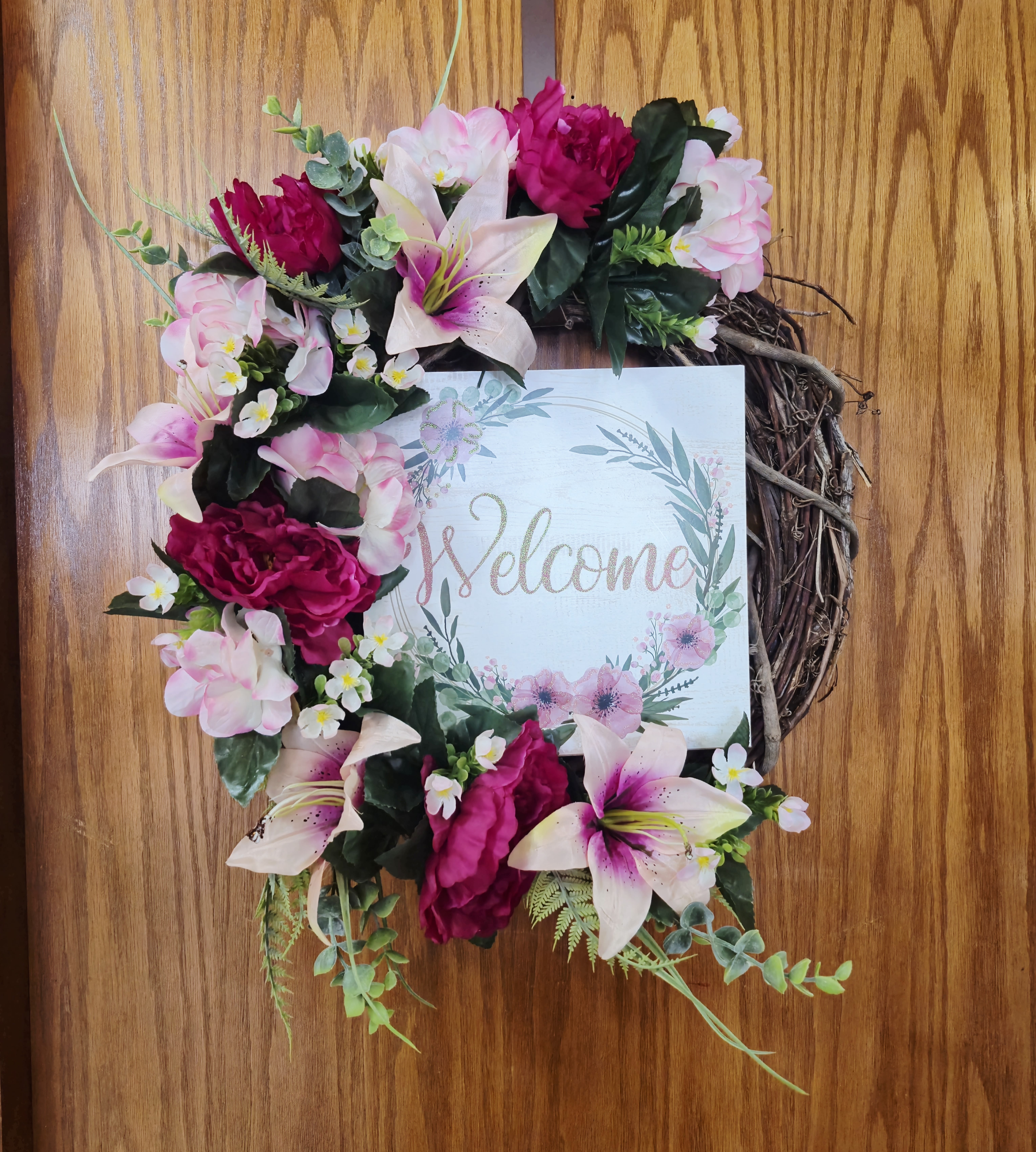 Welcome 18" Grapevine Wreath  - Soft pale pink lilies and rich magenta blooms tumble across a rustic wooden grapevine frame for a cheerful entryway accent - perfect as a welcome wreath for housewarmings or everyday front-door décor. This 18" grapevine wreath arranges silk flowers and mixed greenery around a square "welcome" sign, creating a warm first impression the person you're gifting will see every time they come home.  The palette mixes pink, white, and green: large pale pink lilies, deep magenta blossoms, and small white filler flowers sit among twig branches and leafy accents for texture and depth. The wooden frame and printed sign give it a handcrafted, homey feel while the silk blooms keep the look fresh without maintenance.  Made to hang on an entry door or interior wall, this wreath sends a friendly, inviting message - a thoughtful choice for new homeowners, hosts, or anyone who loves cottage-style decor. The 18" diameter fits most standard doors.