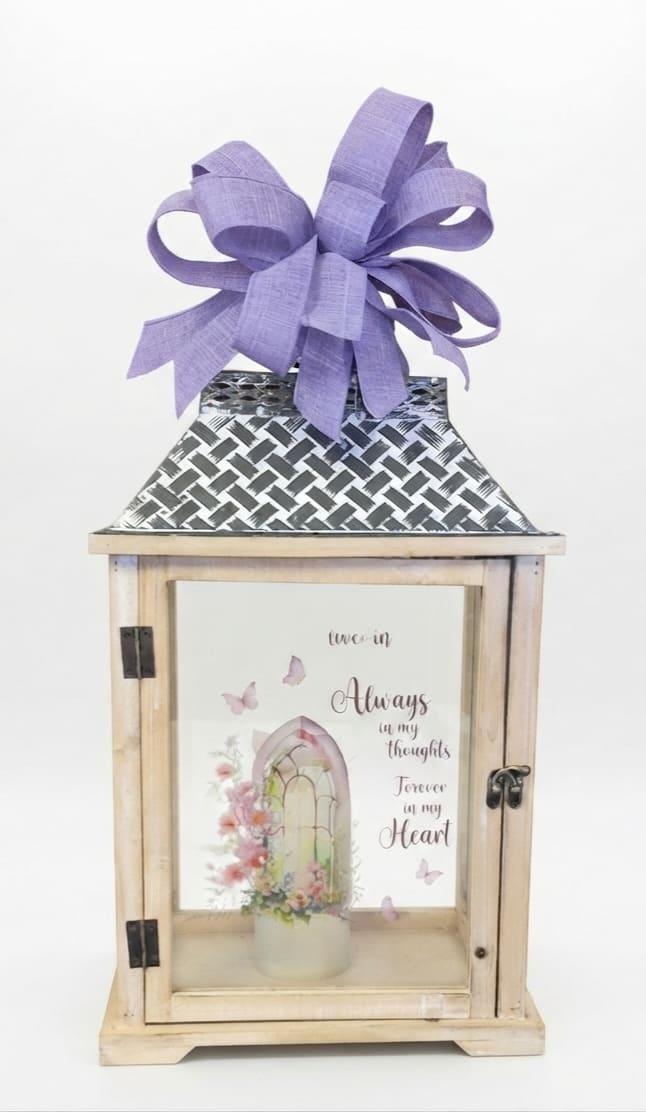 Always in my Thoughts Forever in my Heart Lighted Lantern - A gently glowing wooden lantern crafted as a heartfelt sympathy gift, the Always in my Thoughts Forever in my Heart lighted lantern is designed to bring comfort and soft illumination to a memorial space. This remembrance lantern suits sympathy and condolence occasions, offering the kind of quiet presence that helps the person you care about feel remembered and honored.  The lantern stands 22" tall and 12" wide with a natural wood frame, glass panels, and battery operated lighted candle. Inside a printed memorial insert features a pastel floral illustration in soft pinks with a comforting message and a large lavender ribbon bow tied at the top for a subtle pop of color. The muted purple, white and pink palette and warm wood tones create a soothing, home-ready piece rather than a fresh floral arrangement.  