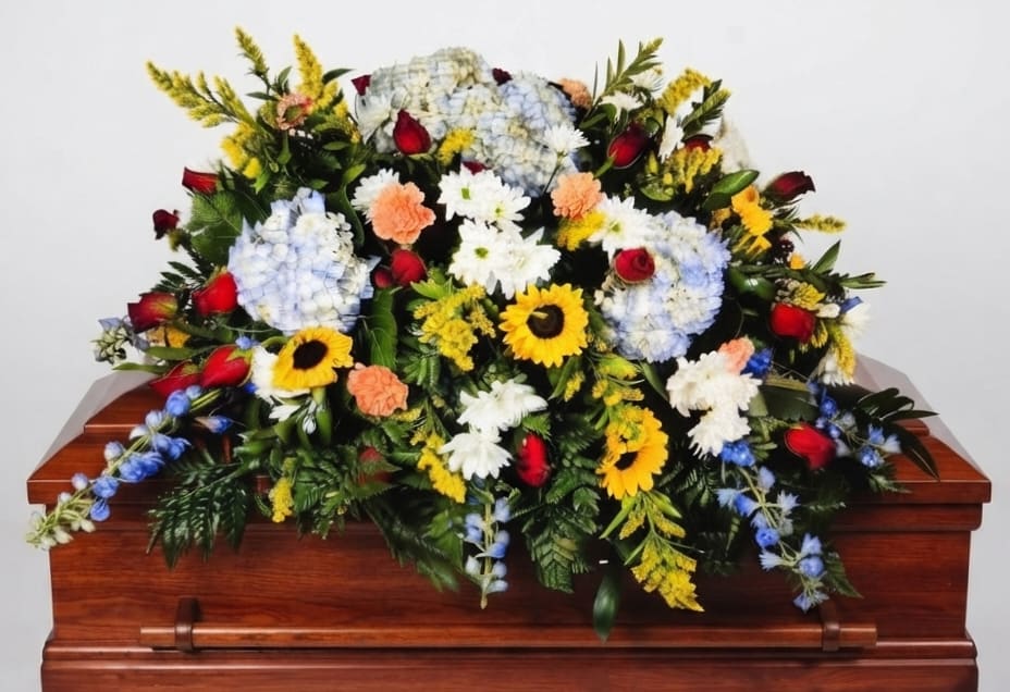 Sunflower &amp; Rose Casket Spray - A broad, cascading casket spray that brings warmth and gentle brightness to a sympathy or funeral service, combining the stately presence of roses with the sunny comfort of sunflowers. This funeral flower arrangement is designed to drape the casket with a traditional, ceremonial silhouette-the kind of piece that helps the person you're honoring feel celebrated and remembered during a difficult moment.  Clusters of pale blue hydrangeas anchor the composition, while deep red roses and white daisies provide classic contrast. Yellow sunflowers and soft peach carnations add uplifting color, and delicate blue delphinium-like blooms and mixed seasonal accents trail into dense greenery for a full, abundant look. The spray is arranged on a casket base for an elegant, flowing presentation that reads well from every angle.  For a meaningful tribute, this arrangement suits memorial services where you want to convey love, warmth, and respect-whether remembering a friend, family member, or cherished colleague. 