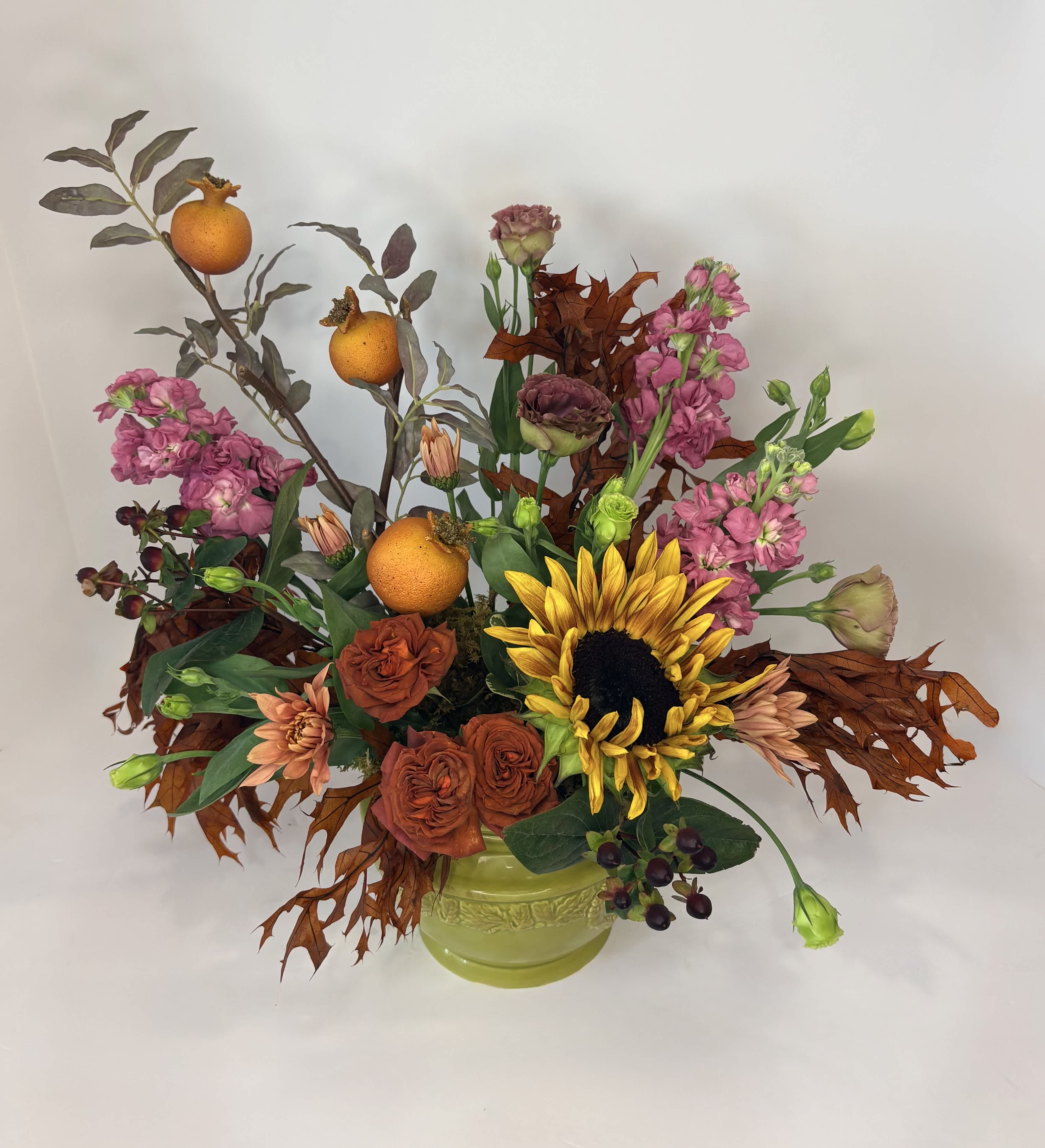 Autumn floral arrangement with sunflowers, roses, pink blooms, and oranges in a green vase