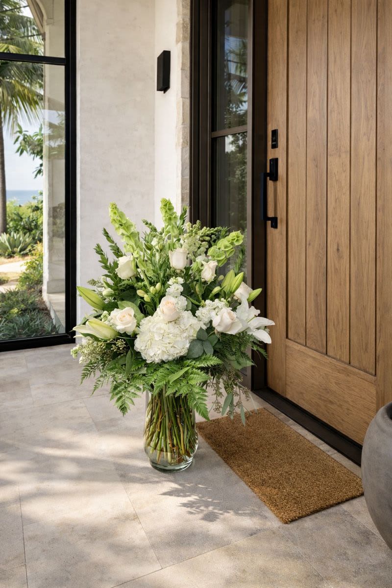 Tall white floral arrangement in a glass vase by a wooden door