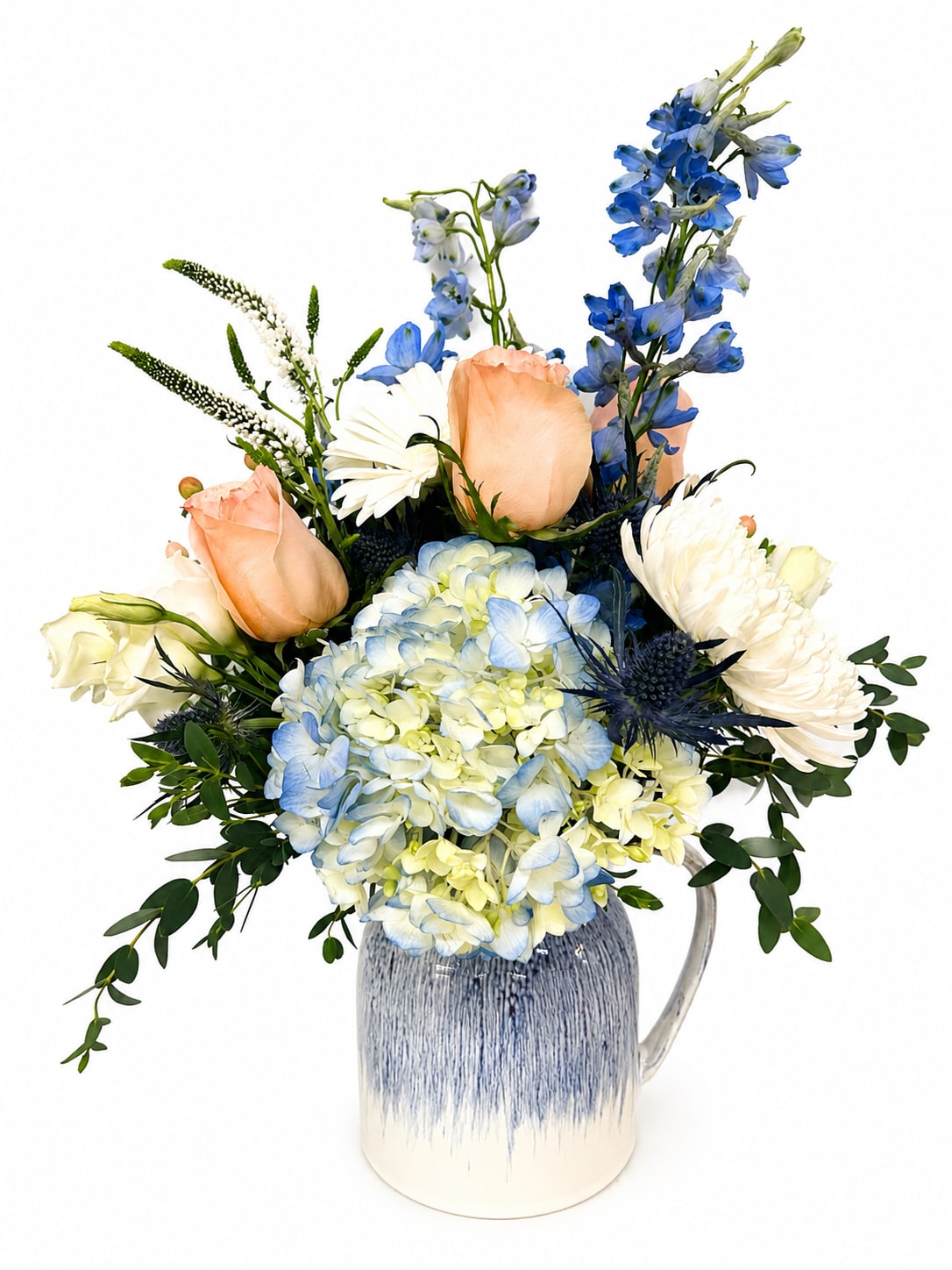 Blue Pitcher Garden - Local Delivery Only  - Fresh, airy, and reminiscent of sunshine on a summer porch, the Blue Pitcher Garden is a charming floral arrangement designed to brighten any day. Perfect as a summer gift, thoughtful surprise, or seasonal centerpiece, this garden-inspired design combines soft peach roses, lush blue-green hydrangeas, crisp white daisy blooms, and striking blue delphinium for a look that feels both relaxed and refined.  Arranged in a beautiful blue-and-white ceramic pitcher vase, the design features full clusters of hydrangeas balanced by the graceful height of delphinium, creating a naturally elegant silhouette. Soft shades of peach, white, green, and blue add depth, texture, and fresh seasonal beauty. The decorative pitcher vase brings a warm, welcoming touch and can be enjoyed long after the flowers have faded.  Whether you're sending a heartfelt "thinking of you," celebrating a special occasion, welcoming a friend, or bringing a hostess gift to a summer gathering, the Blue Pitcher Garden makes every moment feel memorable. Delivered fully arranged and ready to display, it's a beautiful way to make someone feel appreciated and celebrated.  For best results, change the water every two days and trim the stems slightly when refreshing the arrangement.  We proudly offer hand-delivered flower arrangements throughout Smyrna, Marietta, Mableton, Austell, Powder Springs, and Atlanta, Georgia, ensuring your thoughtful gift arrives fresh, beautiful, and ready to enjoy
