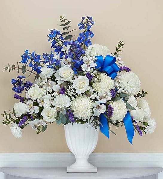 Tall white and blue sympathy flower arrangement in a white pedestal vase with a blue bow
