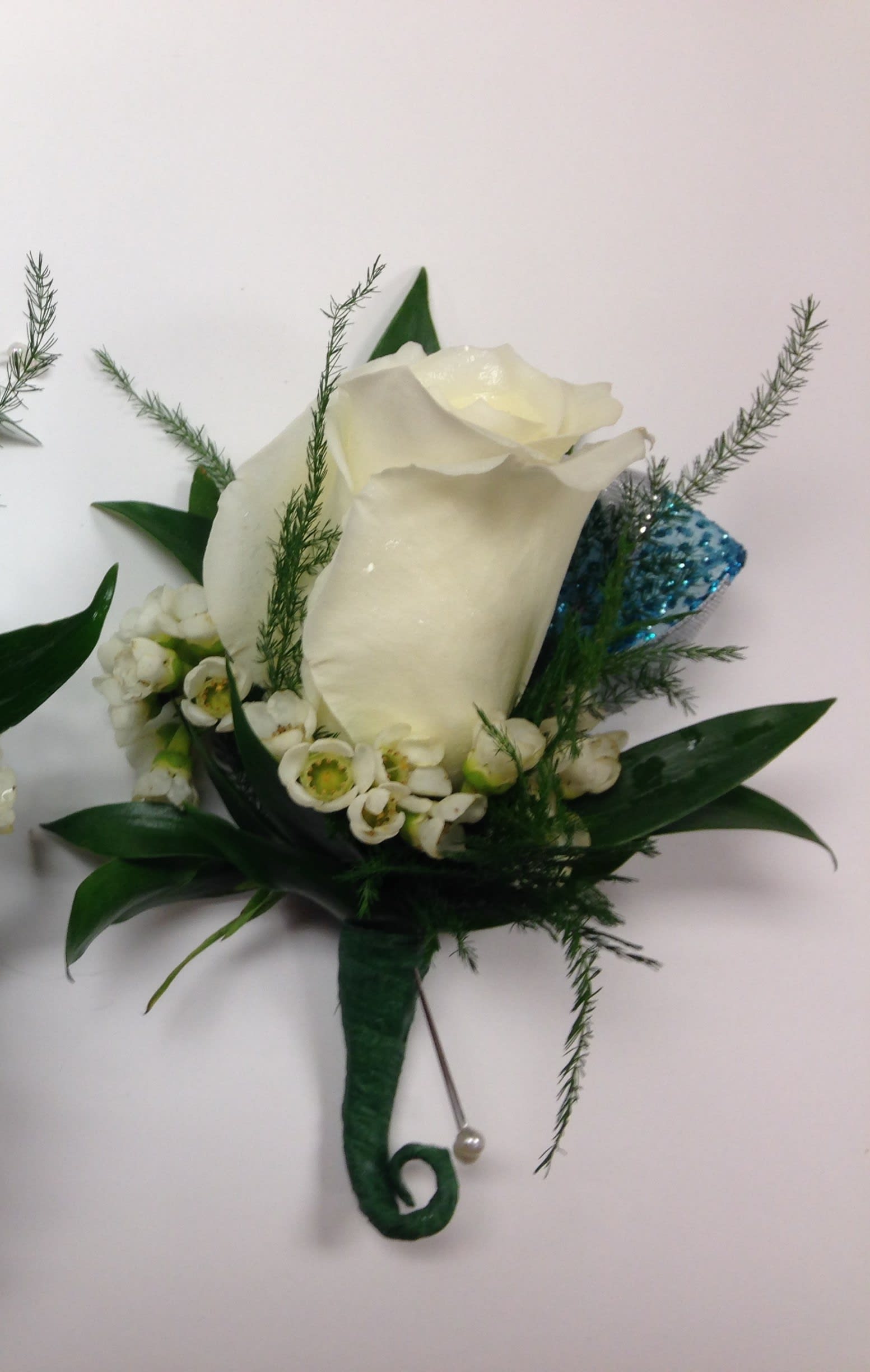 White rose boutonniere with small white blossoms, greenery, and a blue glitter accent wrapped with a pearl pin