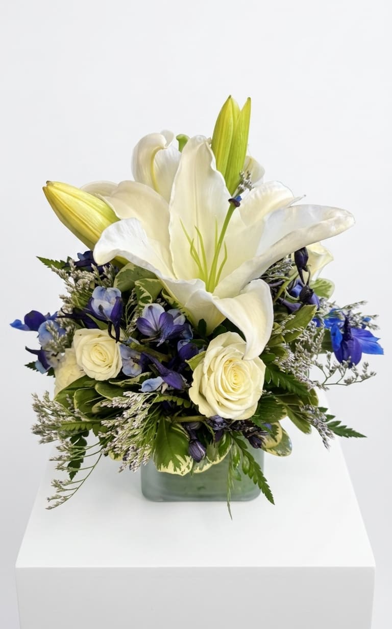 Midnight Moon Bouquet - Capture the serene beauty of a starlit night. This elegant arrangement features a magnificent, large white Oriental lily as its centerpiece, beautifully surrounded by crisp white roses and deep, velvety blue delphinium. Accented with delicate filler flowers and lush, textured greenery in a modern clear glass cube, Midnight Moon Bouquet offers a sophisticated contrast that brings a sense of calm and luxury to any space. Perfect for anniversaries, expressions of sympathy, or a striking centerpiece.
