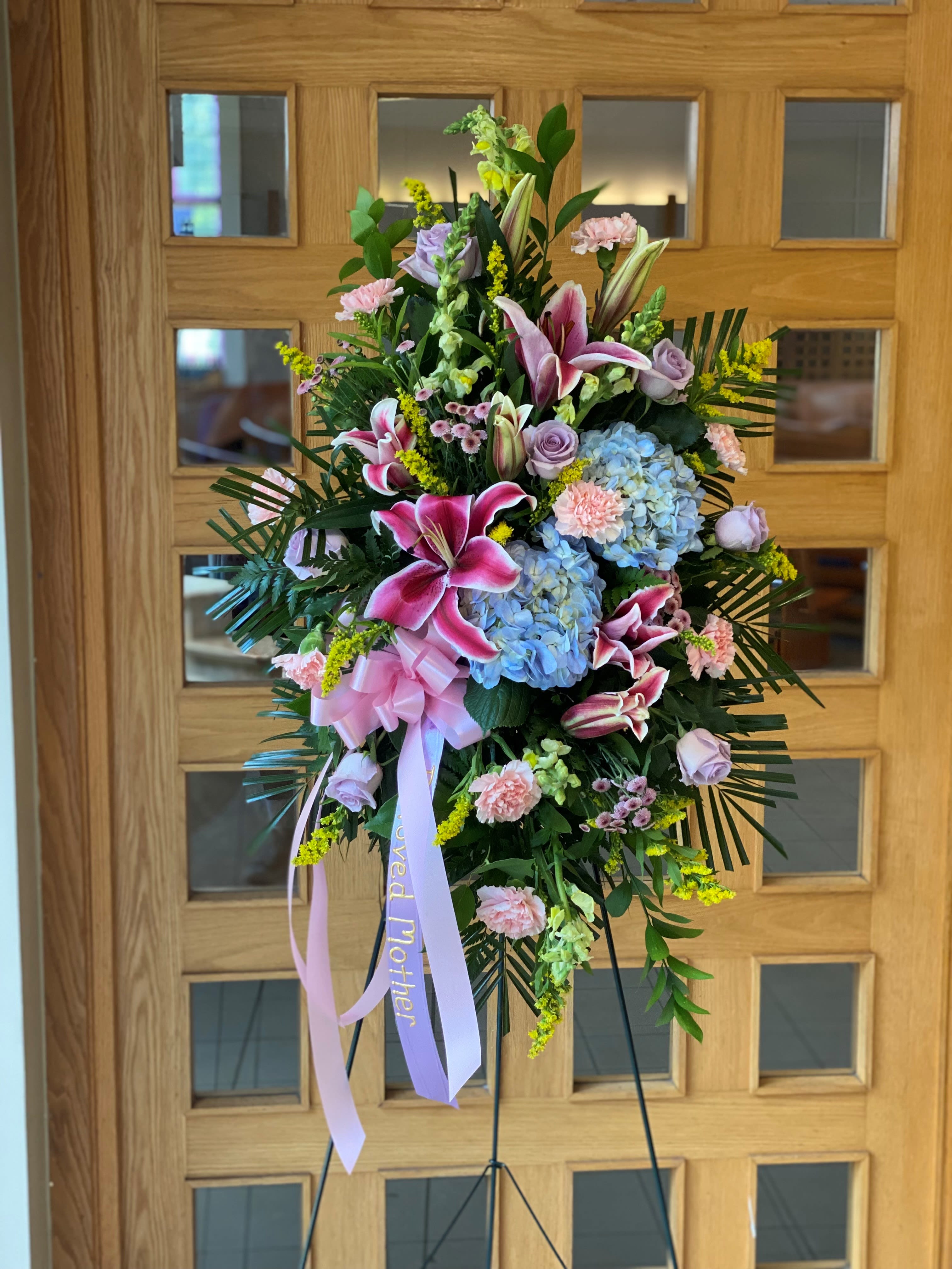 Heaven's Embrace - The combination of stargazer lilies, blue hydrangeas, pink roses and carnations, with touches of yellow solidago and lush greenery, creates a soft yet vibrant tribute. The pink bow and ribbon reading “Beloved Mother” add an extra layer of tenderness, making this arrangement a heartfelt expression of love and remembrance.