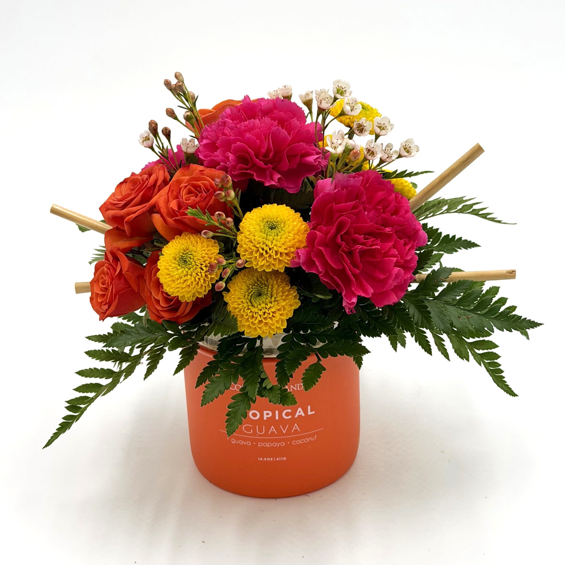 Tropical Sunset Glow - Bright and playful, this arrangement pairs vibrant orange roses with hot pink carnations and cheerful yellow button mums for a bold tropical-inspired look. Designed in a reusable Tropical Guava candle vessel, it brings a fun pop of color and fragrance to any space. Textural greenery and delicate wax flower accents add a fresh garden feel that makes this piece both modern and joyful.