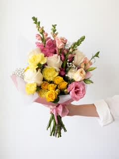 Handheld bouquet of pink, yellow, and white flowers wrapped in pale pink paper