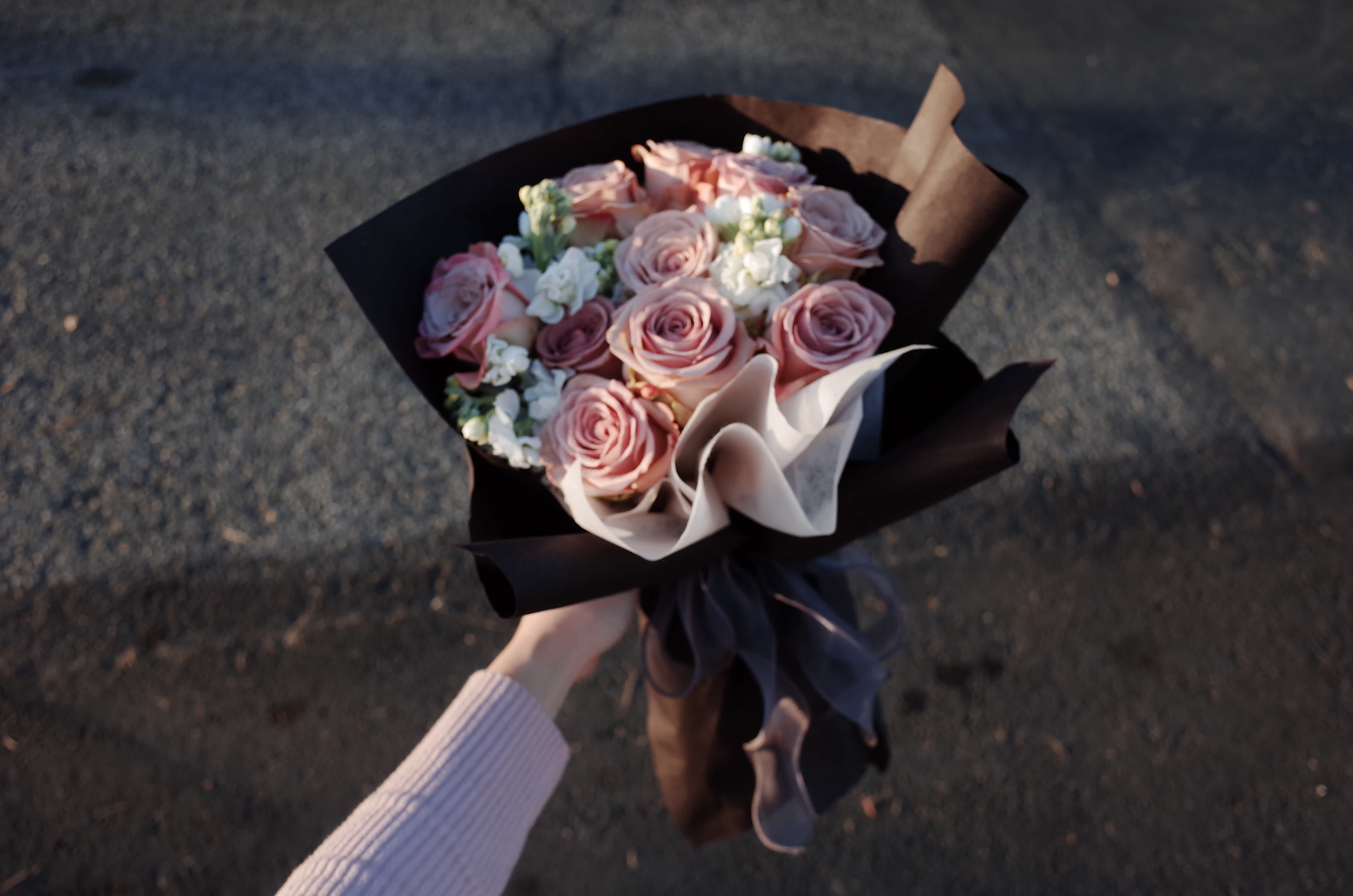 Handheld bouquet of pink roses with white filler flowers wrapped in dark paper
