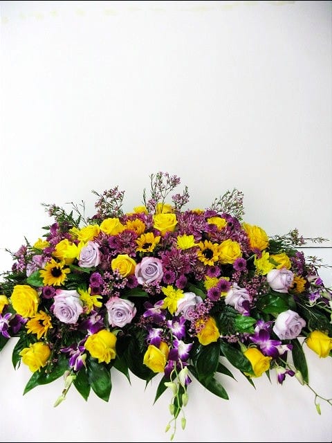 Elongated arrangement of yellow and lavender roses with purple orchids and daisies.