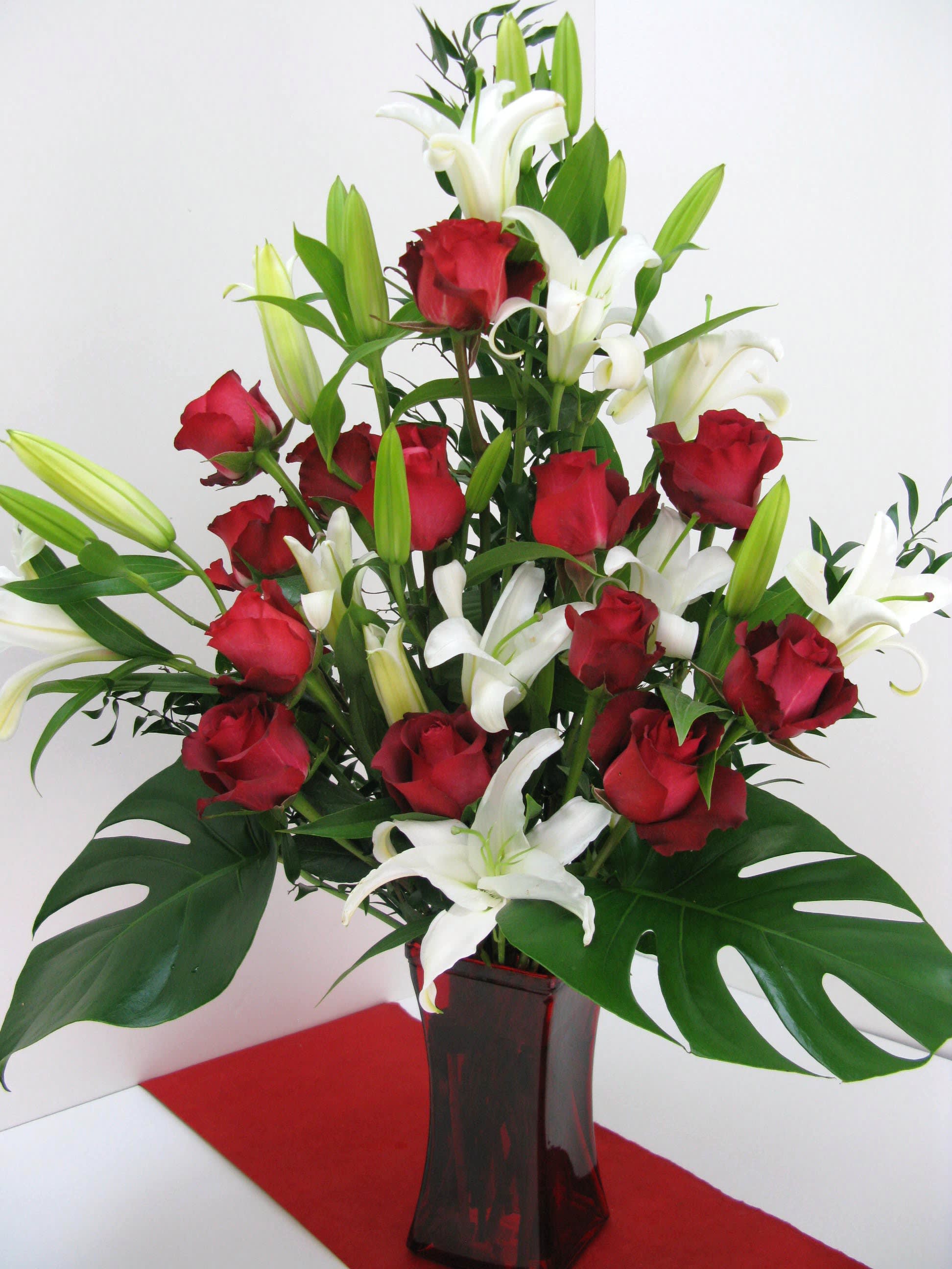 Tall arrangement of red roses and white lilies with large tropical leaves in a red glass vase.