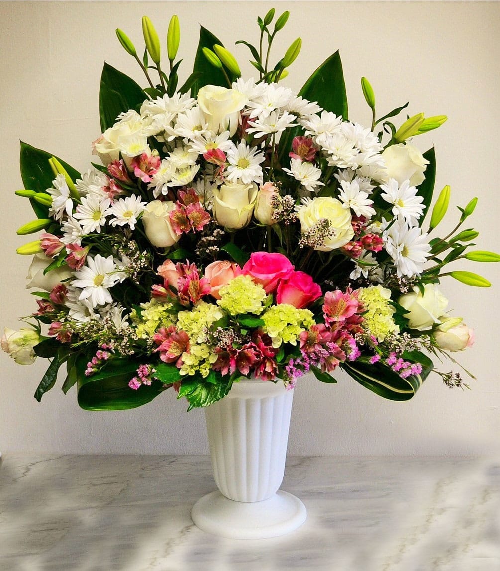 Tall mixed arrangement of white daisies, roses, lilies and pink flowers in a white pedestal vase