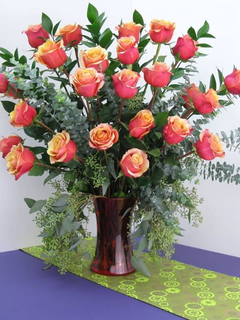 Tall arrangement of pink and yellow roses with lush foliage in a red glass vase