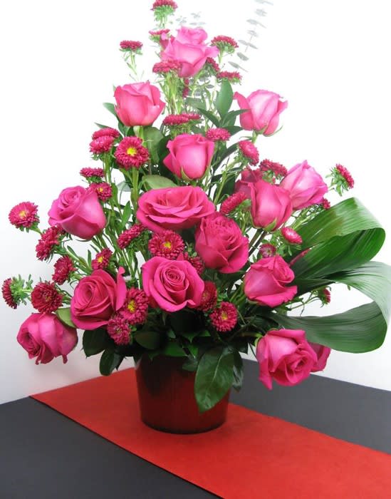 Tall arrangement of hot pink roses and small pink asters in a red pot on a red table runner