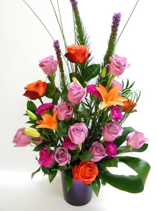Tall arrangement of lavender roses, orange lilies, and fuchsia blooms in a purple vase with a folded green leaf.