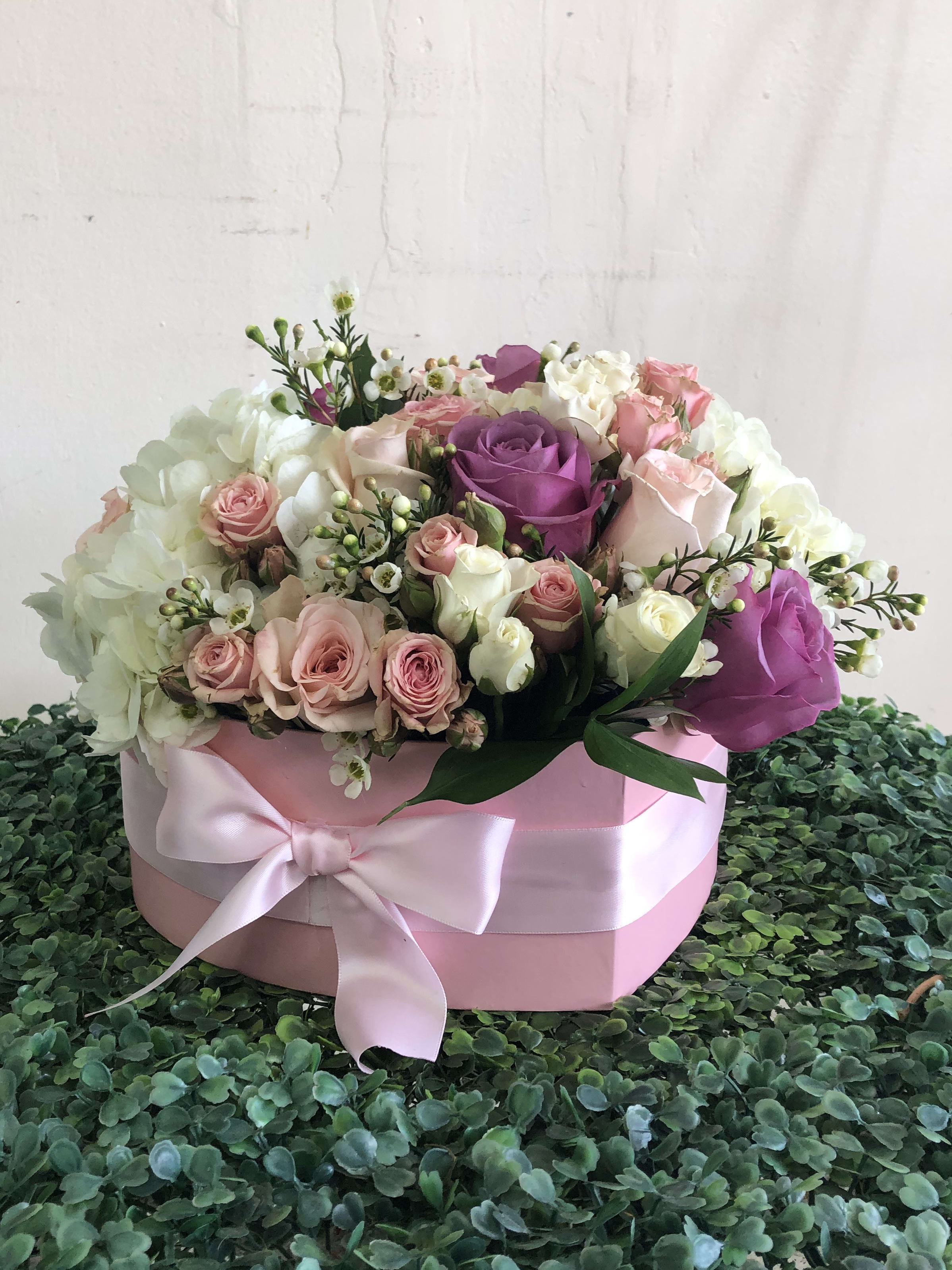 Round pink hatbox of white, blush, and lavender roses with a satin bow on a green textured surface