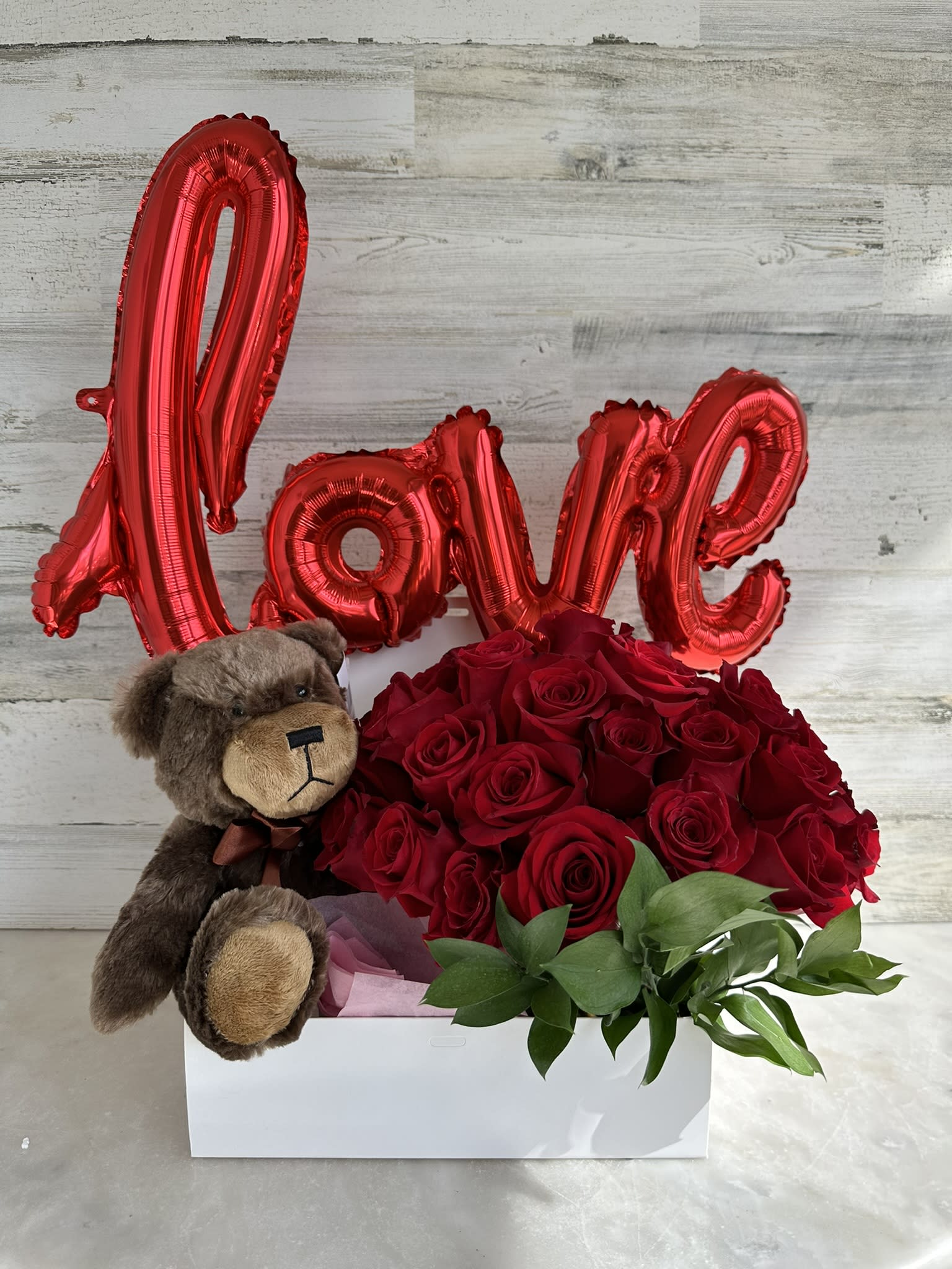 Red roses in a white box with a teddy bear and red "Love" balloon