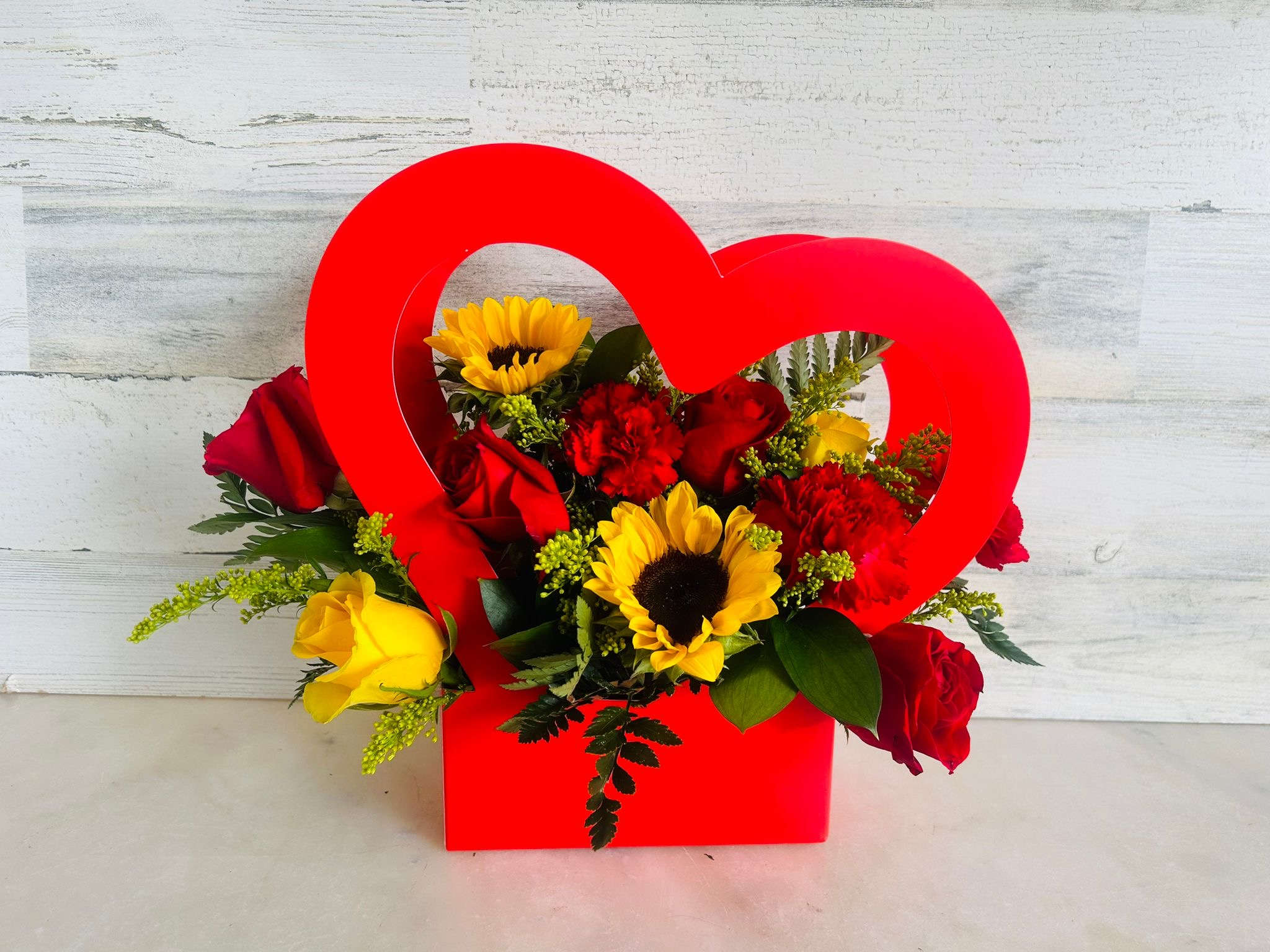 Red heart-shaped box filled with roses, sunflowers, and carnations