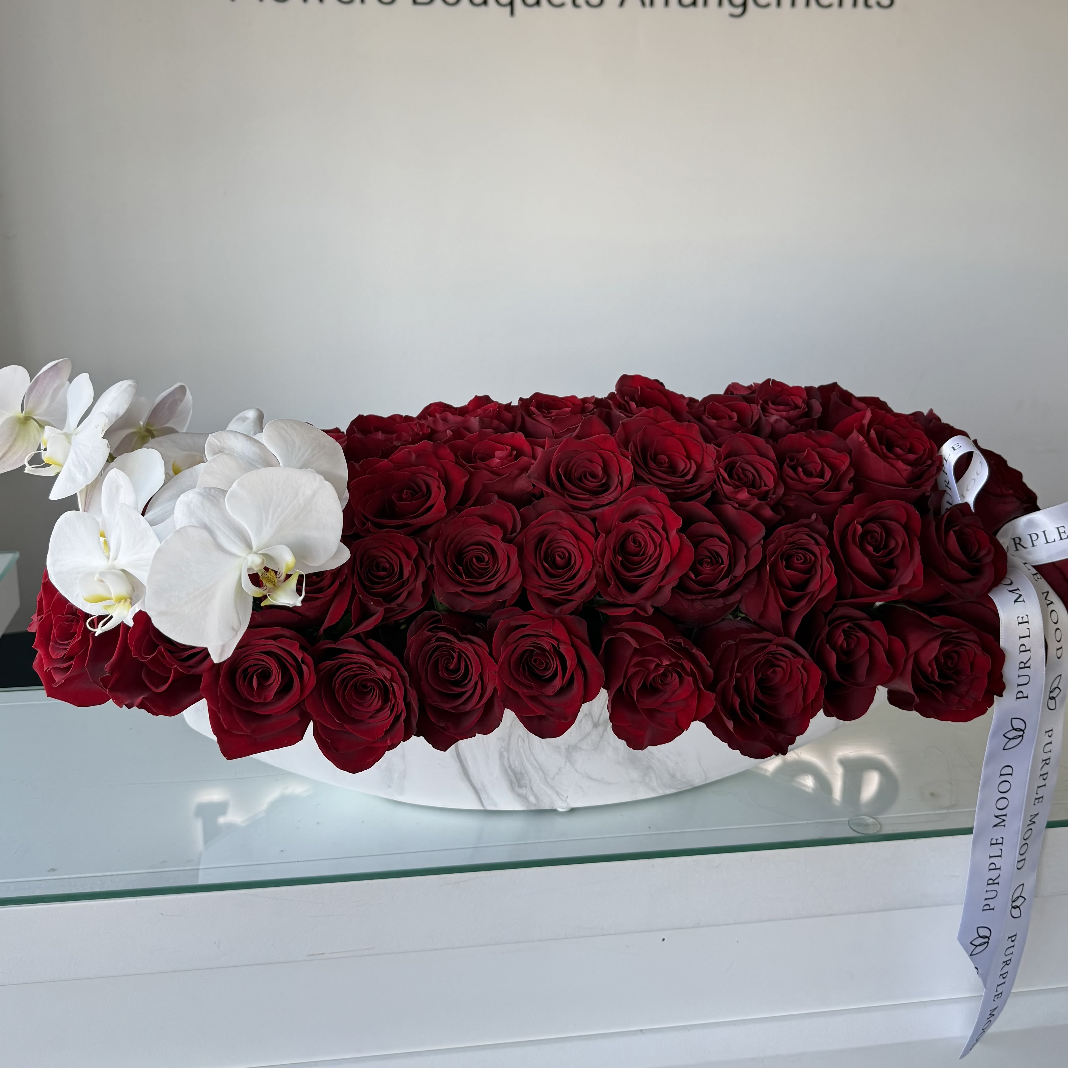 Royal Passion Vase - A luxurious floral statement that embodies timeless romance and sophistication. Featuring a lush bed of deep red roses meticulously arranged in a sleek white marble-textured vase, this piece is gracefully accented with pristine white orchids for a modern contrast. Finished with a signature Purple Mood ribbon, the Royal Passion Vase captures both elegance and intensity — a perfect gift for grand gestures, anniversaries, or luxury interiors.