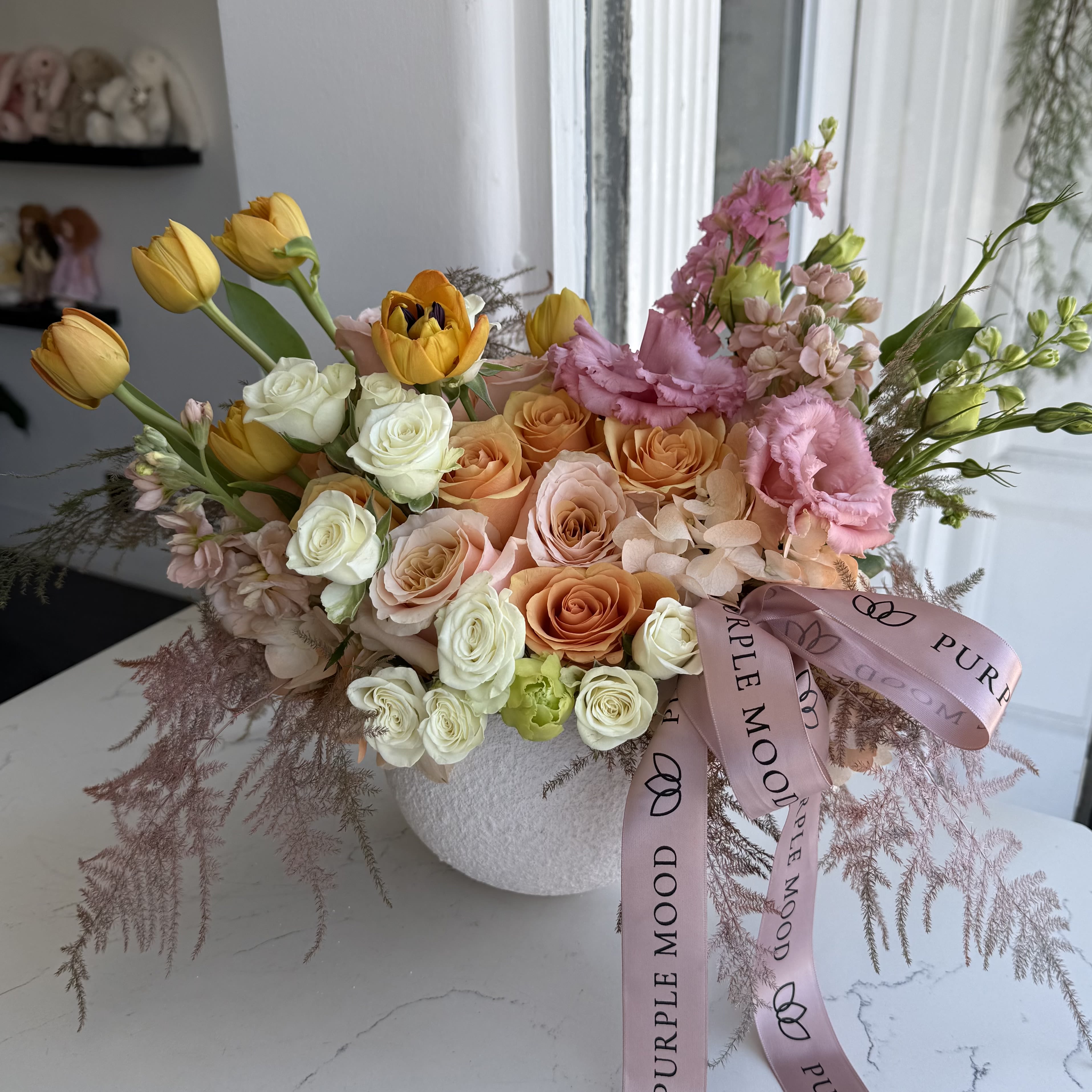 Apricot Dream Vase - Soft, romantic, and effortlessly elegant, the Apricot Dream Vase is a stunning blend of warm peach, ivory, and blush tones. Featuring lush garden roses, delicate tulips, fragrant freesia, and airy seasonal foliage, this arrangement brings a dreamy, ethereal touch to any space. Designed in a chic textured vase and adorned with a graceful ribbon, it’s the perfect choice for celebrating love, milestones, or simply brightening someone’s day.