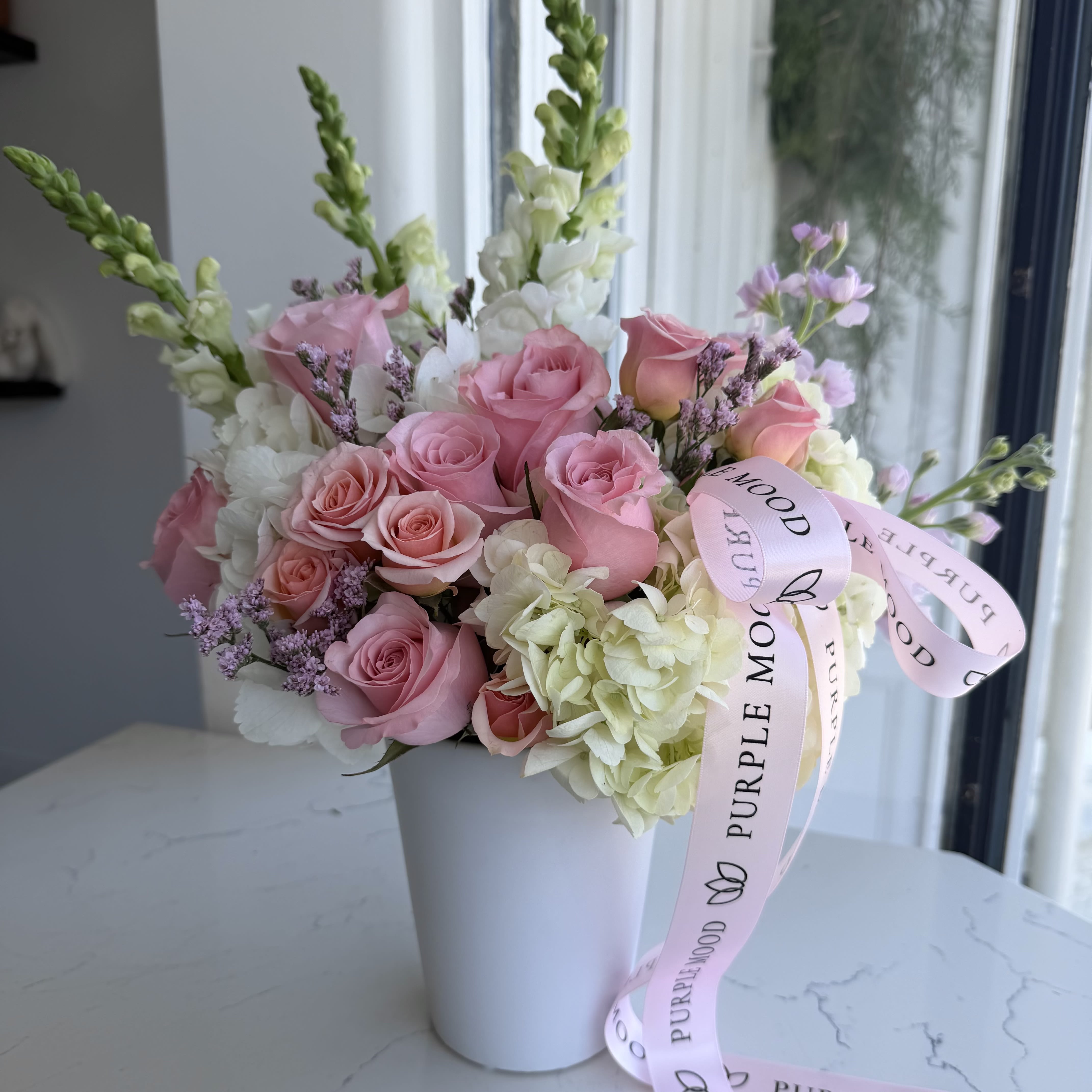 Enchanted Garden Vase - A soft pastel dream captured in a vase — romantic, airy, and full of charm. Shades of blush, lavender, and cream come together in a perfectly balanced design that feels both elegant and effortless. Finished with a satin ribbon, this arrangement brings a sense of calm beauty to any space or occasion.      •    Handcrafted in gentle pastel tones     •    Presented in a modern white vase     •    Finished with a soft satin ribbon     •    Perfect for birthdays, celebrations, or thoughtful gestures
