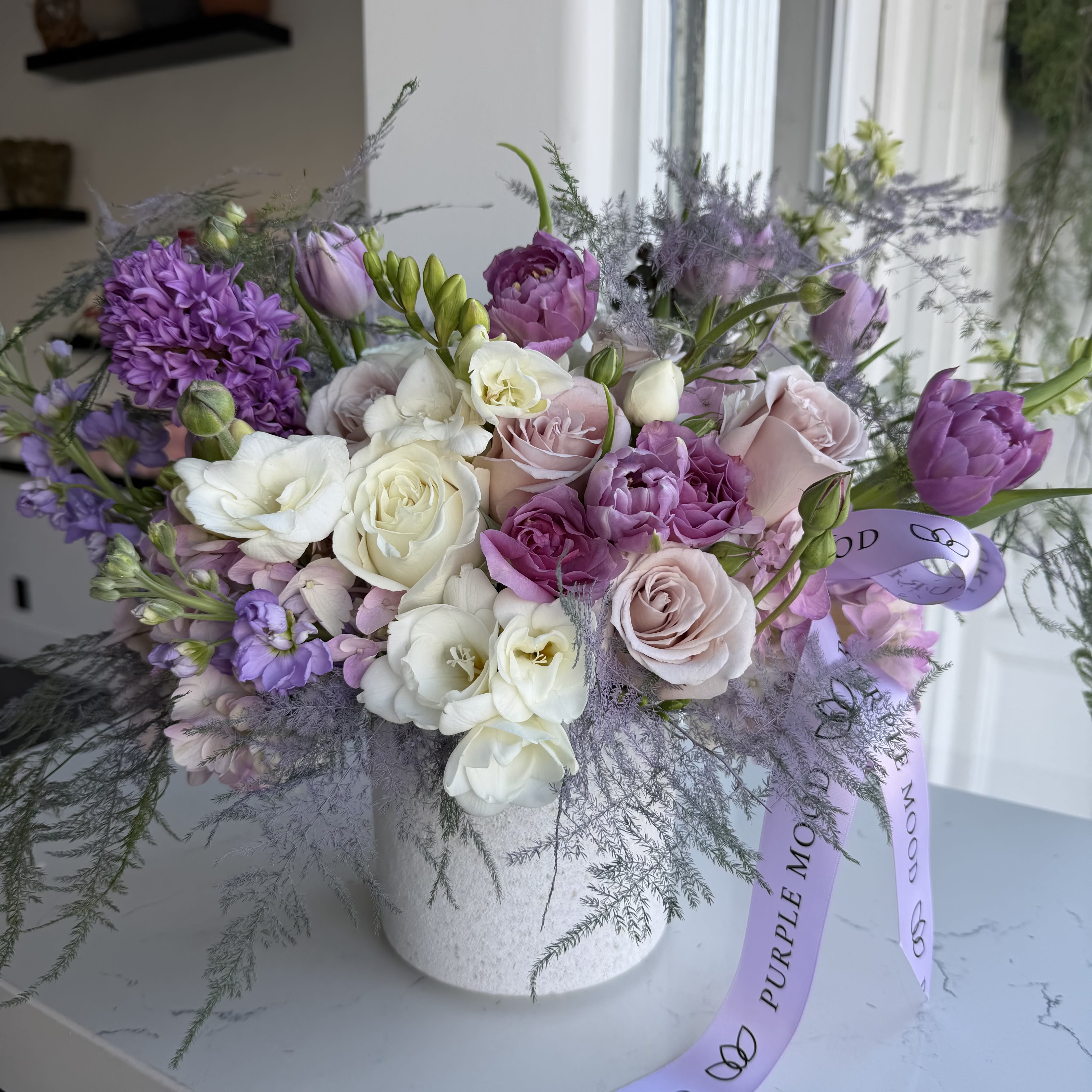 Lavender Bliss Vase - A dreamy arrangement of soft lavender, creamy ivory, and delicate blush roses paired with tulips, dahlias, and lush lisianthus. Accented with airy foliage and artfully arranged in a textured white vase, this bouquet exudes elegance and romance — perfect for brightening any space or celebrating life’s special moments.