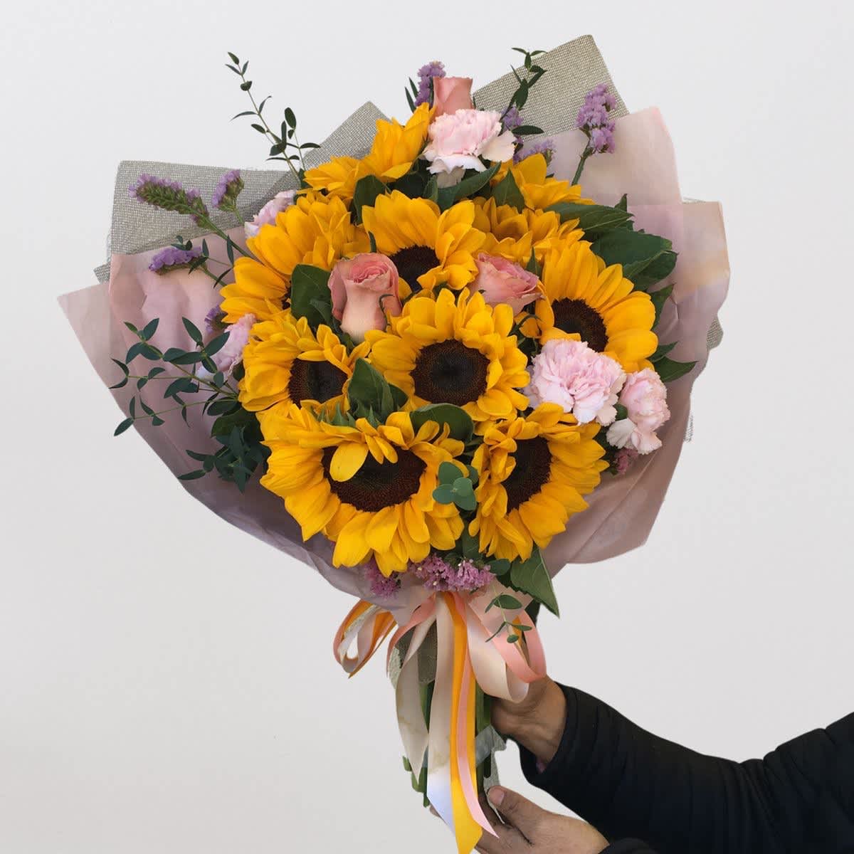 Handheld bouquet of sunflowers, pink roses, and carnations wrapped in pastel paper