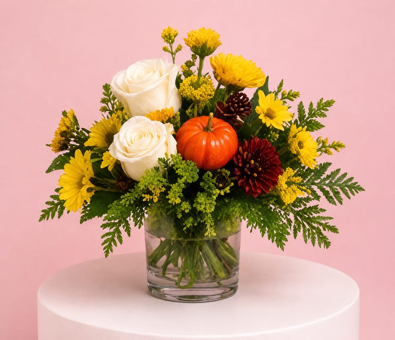 White roses and yellow flowers arranged in a glass vase with a small pumpkin