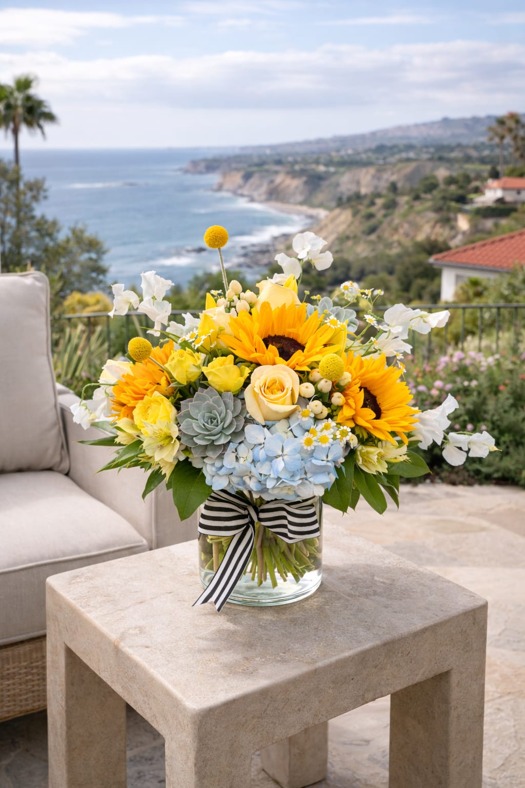 Sunshine on the Coast - Inspired by the golden light and coastal beauty of Palos Verdes, Sunshine on the Coast is a radiant, garden-style arrangement designed to feel both effortless and artfully composed. Warm sunflowers and soft crème de la crème roses take center stage, layered with airy textures and unexpected details for a fresh, modern coastal feel.  Blue hydrangea adds a cool ocean-toned base, while yellow spray roses, tulips, and alstroemeria bring soft movement and warmth throughout. Succulents offer a sculptural, earthy touch, complemented by delicate chamomile, playful billy balls, and whimsical sweet peas. Finished with coffee berry for natural texture, this arrangement feels like a sunlit moment overlooking the coast.  Thoughtfully designed with a balanced mix of color, texture, and form, this piece is perfect for Mother’s Day or anyone who deserves something bright, beautiful, and a little transportive.  Includes: blue hydrangea, sunflowers, crème de la crème roses, yellow spray roses, succulents, alstroemeria, coffee berry, chamomile, billy balls, sweet peas, and tulips.