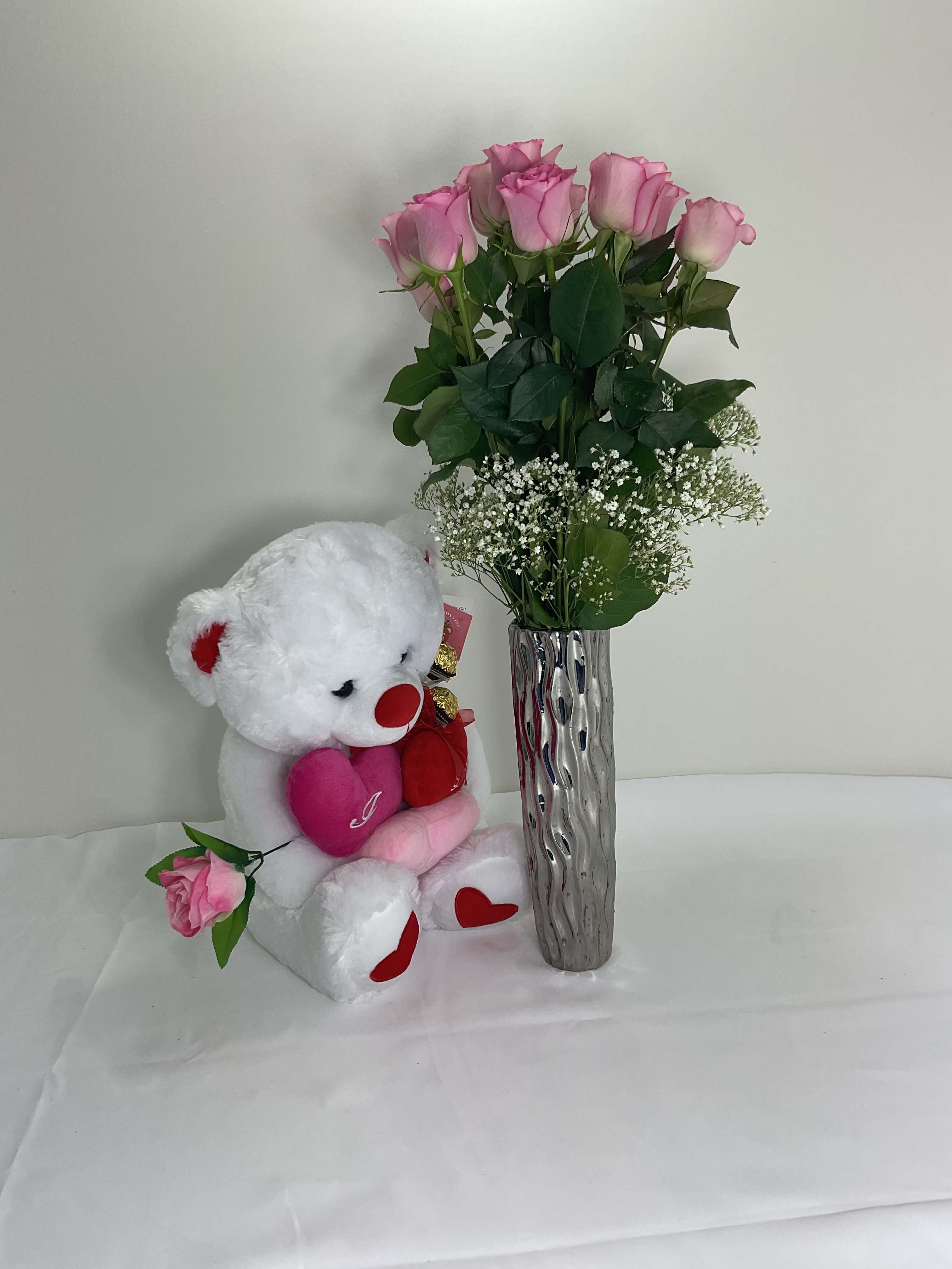 Pink roses in a tall silver vase beside a white teddy bear holding hearts