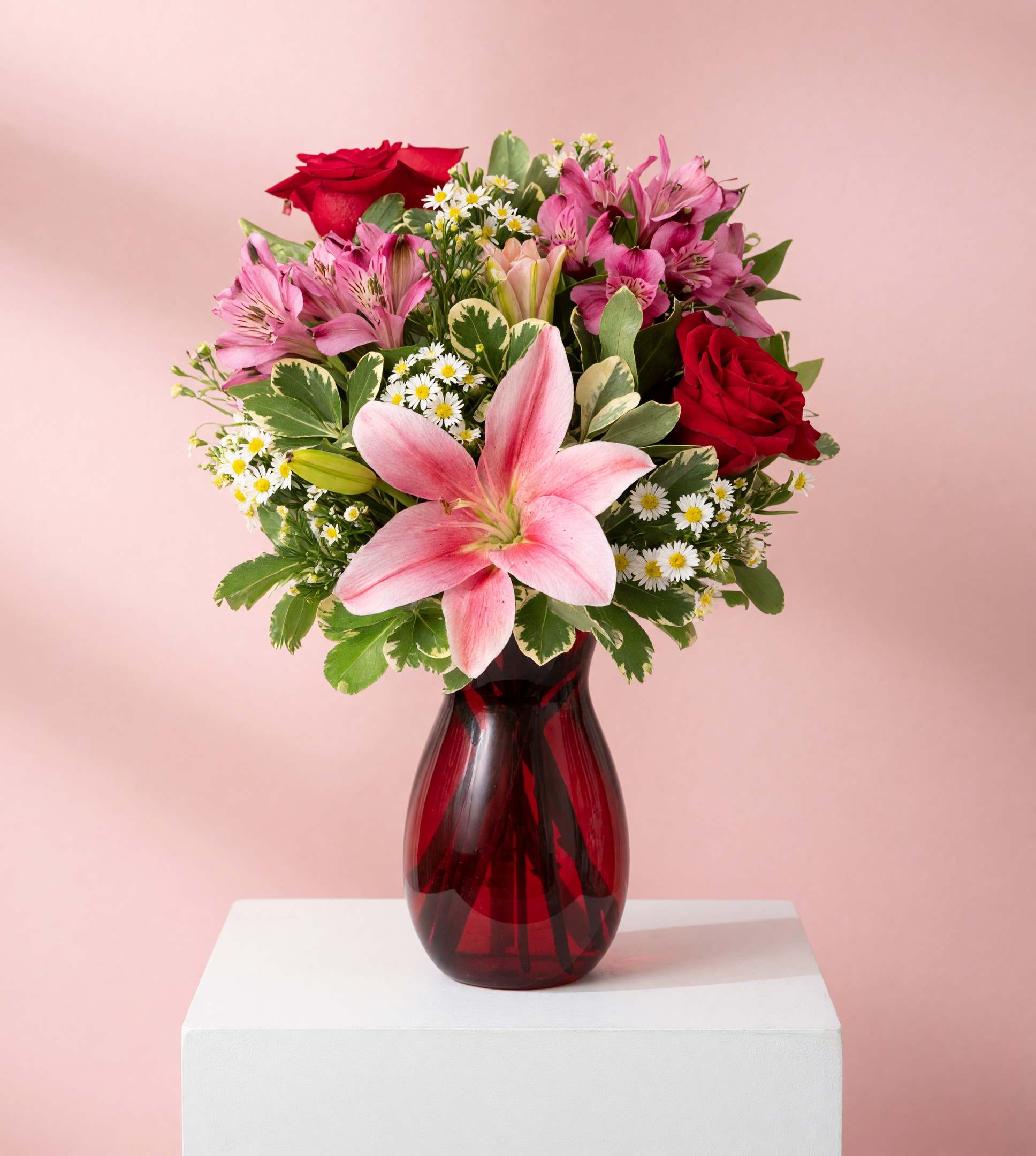 Bouquet of pink lilies, red roses, and small white daisies in a red glass vase
