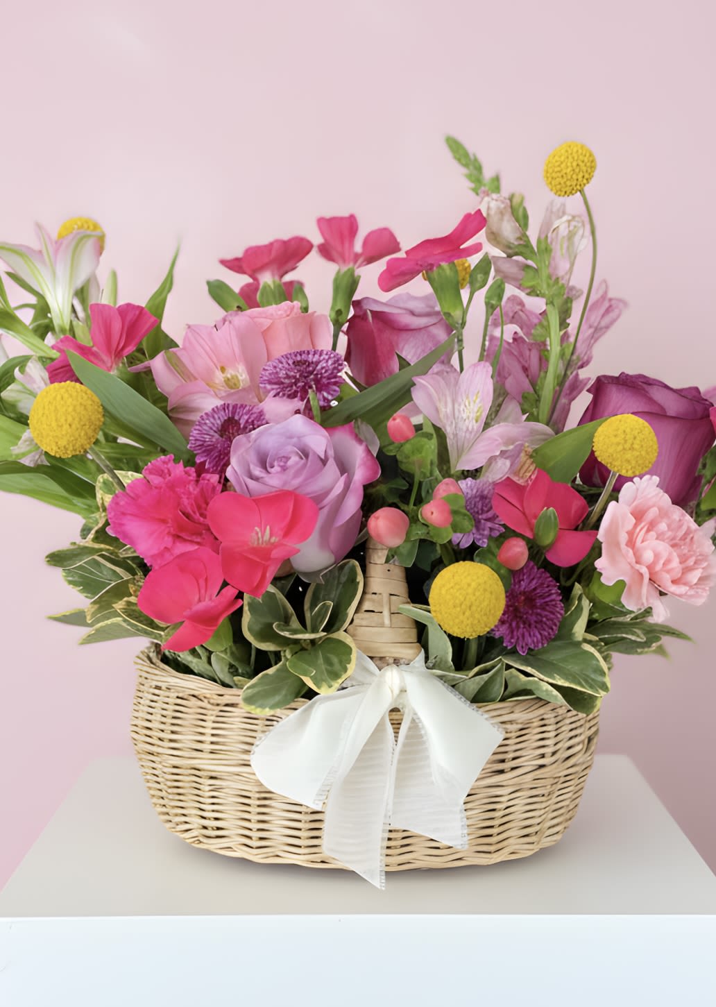 Wicker basket of pink, purple, and yellow mixed flowers with a white bow.