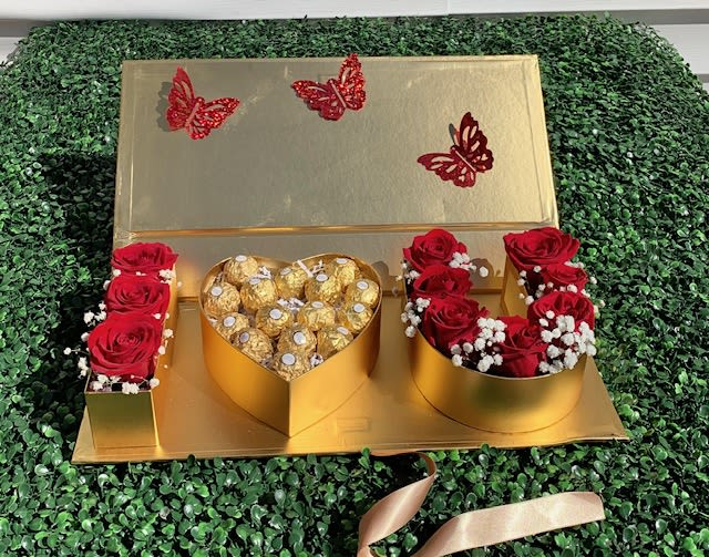 The gold Standard - Imagine a gleaming gold gift box, its surface smooth and reflective like a treasure chest, catching the light with every tilt. The lid is slightly raised or secured with a rich satin ribbon—perhaps crimson or champagne—hinting at the luxurious delight inside.  Open it, and you're met with a stunning contrast: lush red roses, their velvety petals unfolding in bold, romantic layers, arranged snugly in one half of the box like a blooming bed of passion. Each rose is perfectly fresh, bursting with color and scent.  Nestled beside them is an array of gourmet chocolates—elegantly nestled in golden or bronze foil. Think melt-in-your-mouth truffles, heart-shaped pralines, or silky chocolate-covered caramels, each one offering a moment of indulgence that mirrors the richness of the roses.