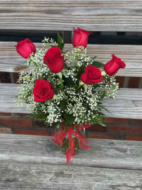 Glass vase of six red roses with white filler flowers and a red bow on a wooden bench