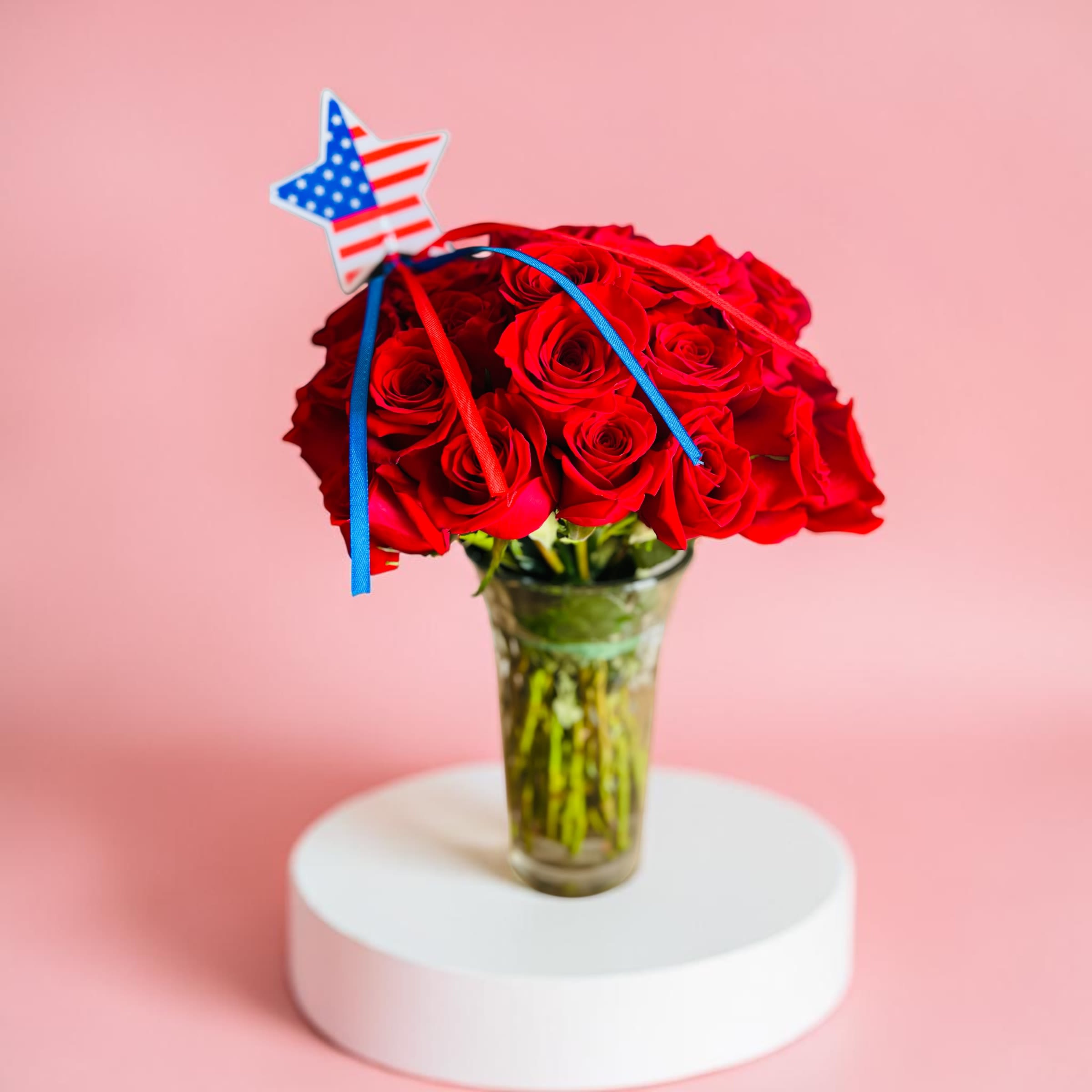 Patriotic Red Roses by Amanda - A lush cluster of vivid red roses arranged with generous green foliage creates an immediate, classic impression-perfect as a birthday bouquet, anniversary roses, or a patriotic gift for Memorial Day and the Fourth of July. This flower arrangement for celebrations blends romantic intent with a subtle nod to national pride: choose the amber glass vase for a warm, vintage feel, or the clear glass option topped with a small star-shaped American flag pick and red-and-blue ribbons for an unmistakable holiday accent. 