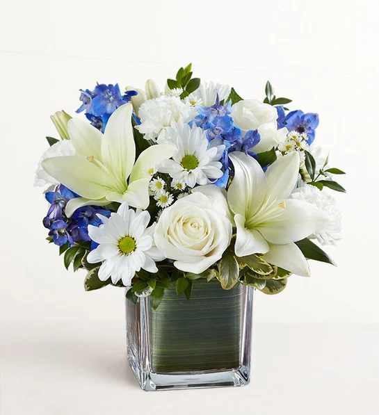 White lilies, roses, and blue flowers in a square glass vase