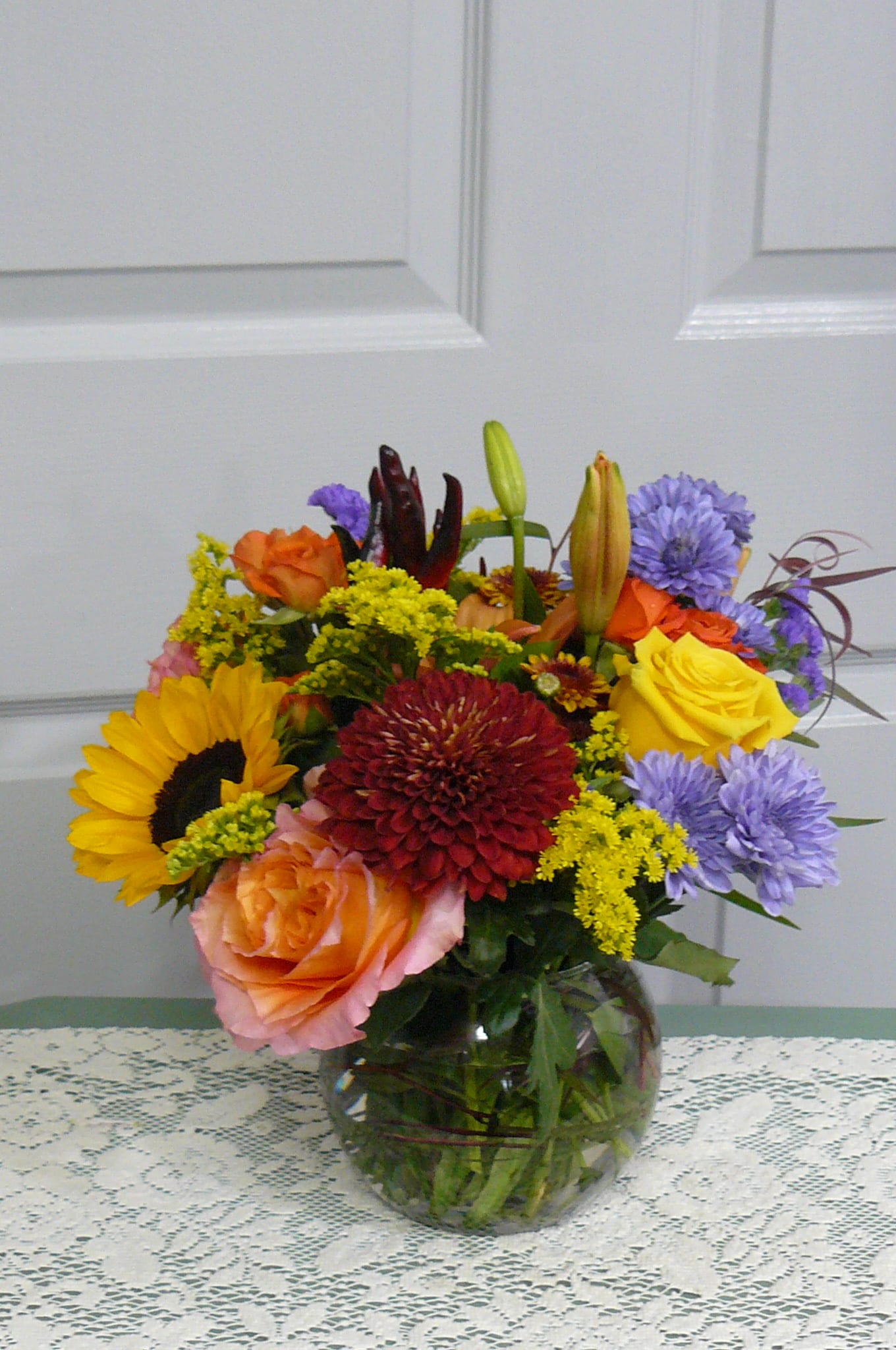 Mixed bouquet in a glass vase with roses, sunflowers, lilies, and purple blooms
