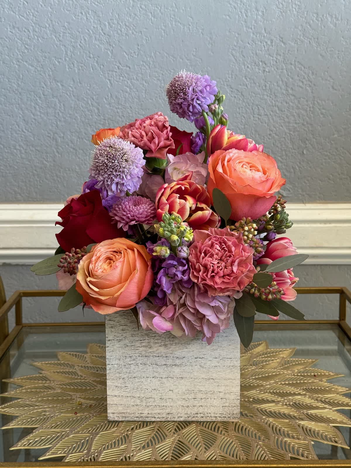 Mixed bouquet of pink, peach, purple, and red flowers in a square vase