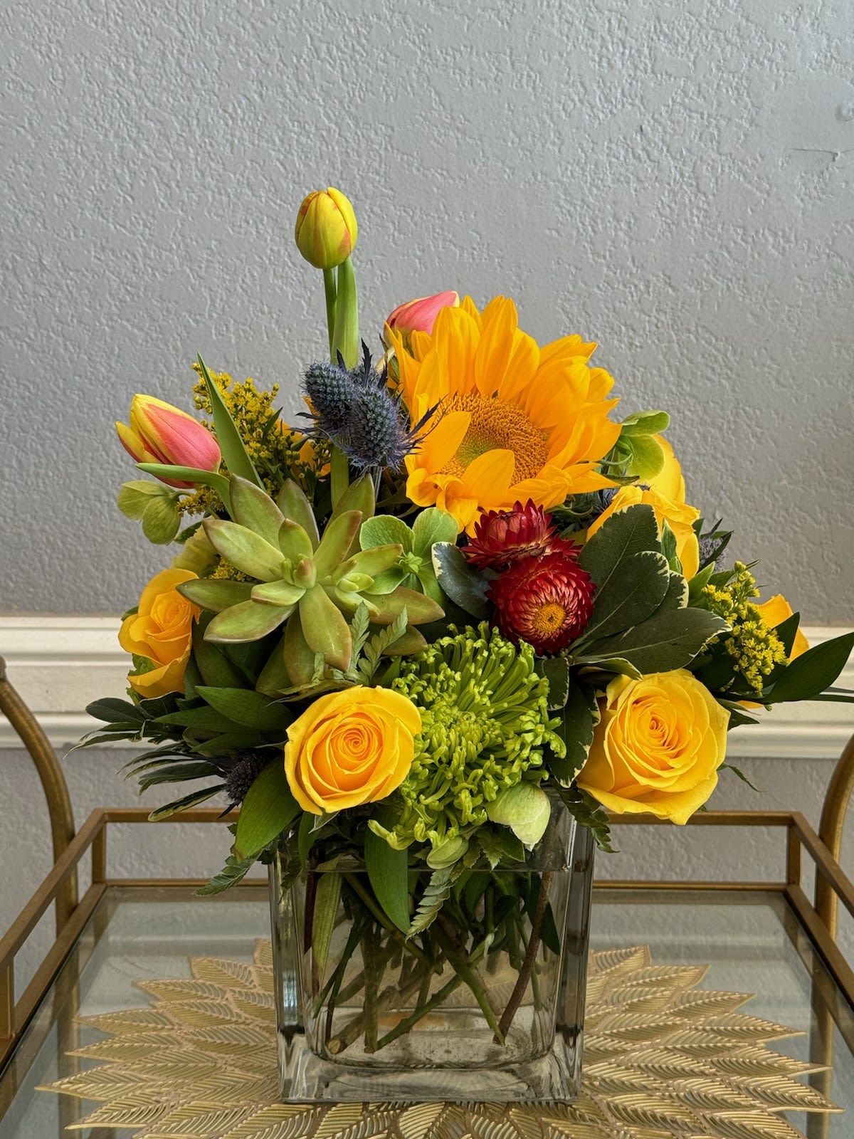 Yellow roses, a sunflower, and succulents arranged in a clear glass vase