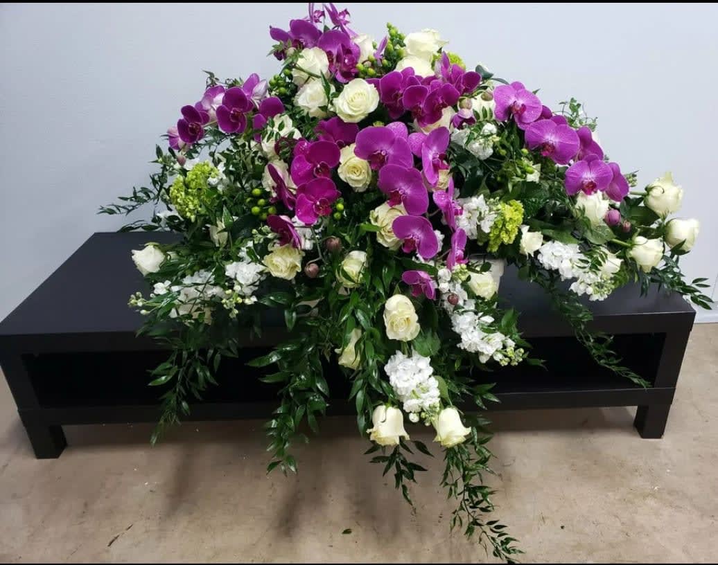 Royal Eternity Casket Spray - This full-size casket spray is a graceful and luxurious tribute, featuring elegant purple Phalaenopsis orchids paired with classic white roses, soft hydrangea, and fragrant stock. Accented with lush Italian ruscus foliage, the arrangement conveys both beauty and reverence. Designed to honor a cherished life, this stunning display offers a heartfelt expression of love, remembrance, and peace.  Please note: Floral variety may vary based on seasonal availability, ensuring each tribute is fresh and unique.