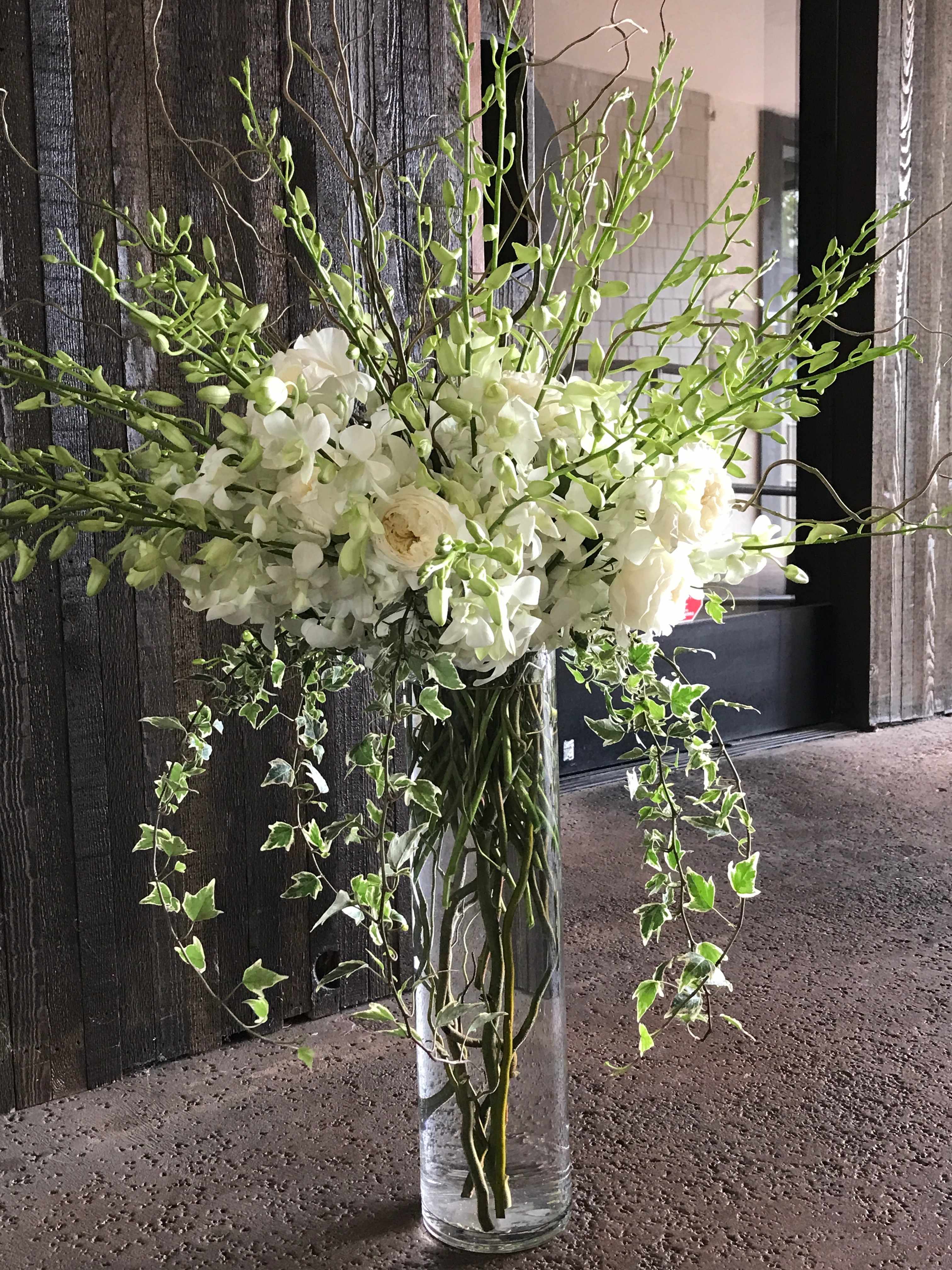 Tall white floral arrangement in a clear glass vase with trailing ivy