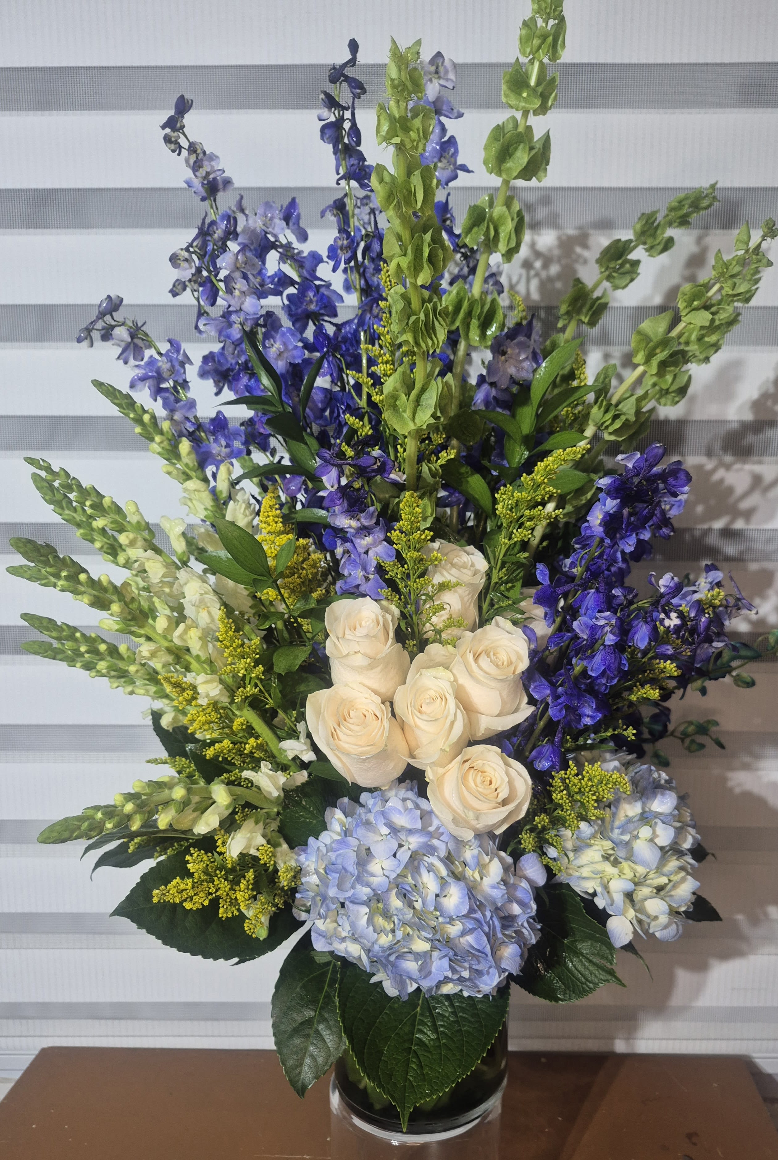 BLUE RHAPSODY - AN ALL AROUND ARRANGEMENT IN AN 11 INS TALL GLASS VASE, CONSISTING OF BLUE DELPHINIUMS, BELLS OF IRELAND, WHITE SNAP-DRAGONS, WHITE ROSES, BLUE HYDRANGIAS AND YELLOW SOLIDAGO FILLER.