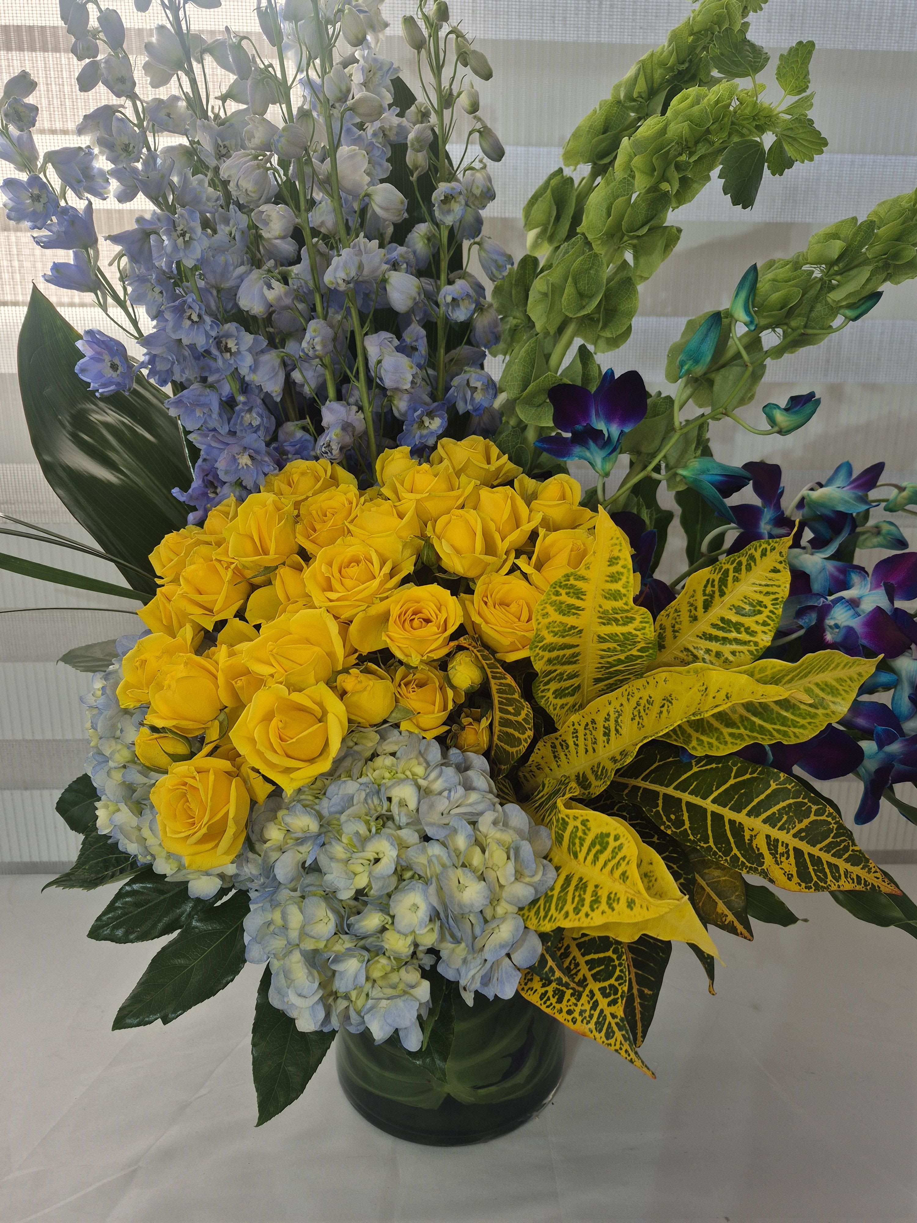 BLUE SKY SUNSET - A ONESIDED ARRANGEMENT MADE INTO A LEAF LINED 10 INCHES TALL GLASS VASE. FLOWERS TO INCLUDE YELLOW GARDEN / SPRAY ROSES, BLUE HYDRANGEAS, BLUE DELPHINIUMS, BELLS OF IRELAND, BLUE/LAVENDER ORCHIDS AND CROTON LEAVES.