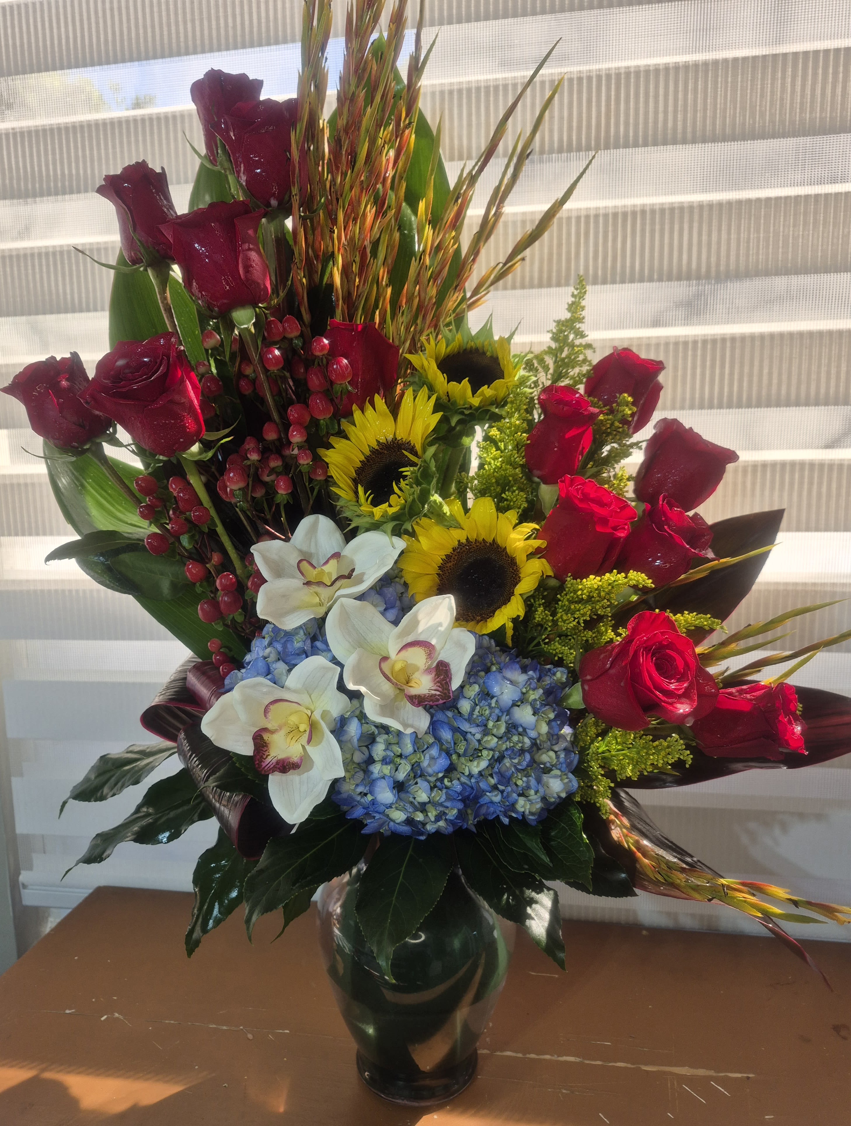 TRUE LOVE ELEGANCE -  A TALL ONE SIDED EXOTIC ARRANGEMENT IN A 11 INCHES TALL GLASS VASE. ARRANGEMENT CONSISTS OF RED ROSES, YELLOW SUNFLOWERS, BLUE HYDRANGEAS, WHITE CYMBIDIUM ORCHIDS BLOOMS, RED/ORANGE HYPERICUM BERRIES, YELLOW SOLIDAGO FILLER AND AND AN ARRAY OF EXOTIC  LEAVES.
