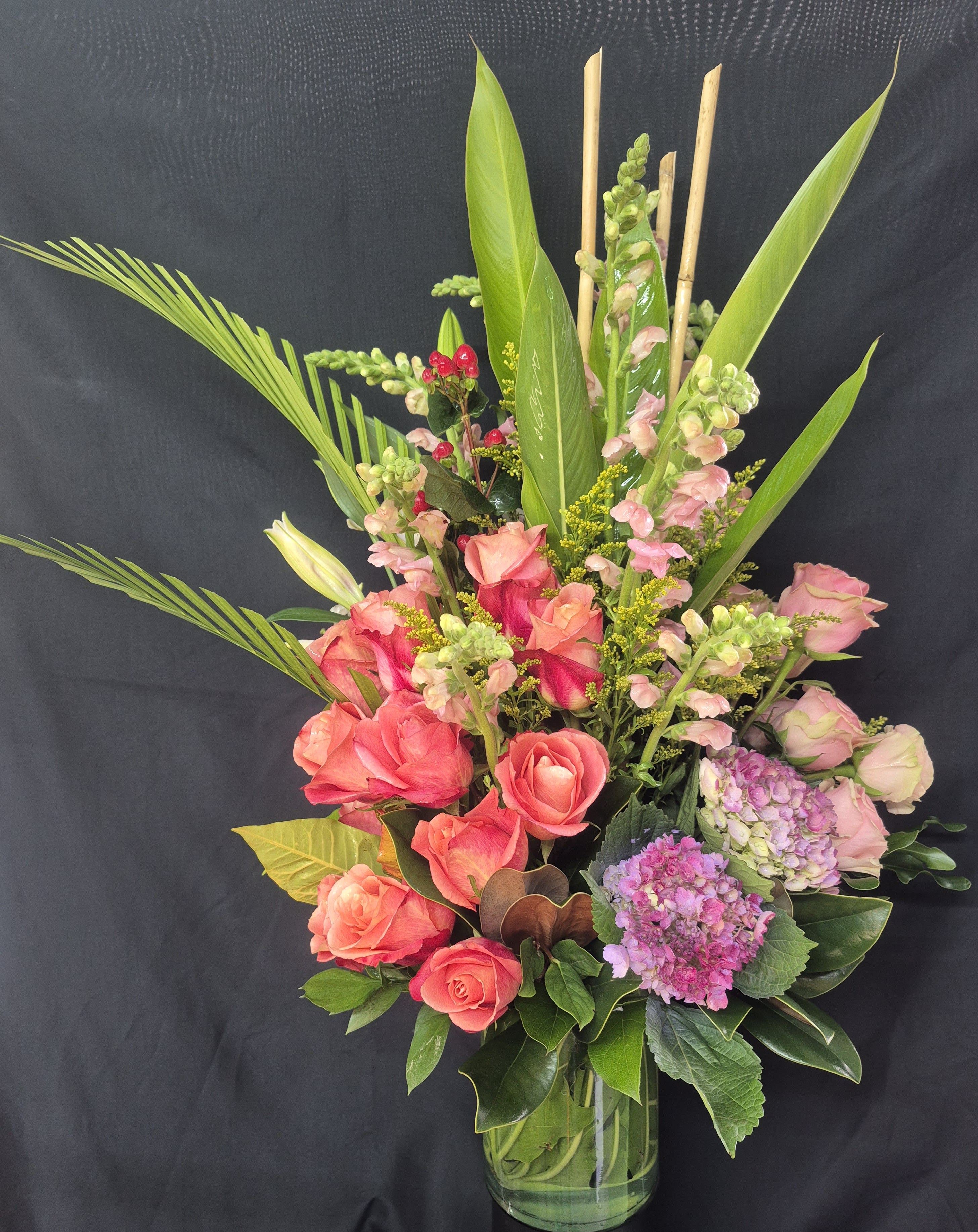 PINK CALYPSO SERENADE - This stunning 360-degree arrangement stands approximately 28-30 ins tall and makes a beautiful statement from every angle. Featuring lush coral and hot pink roses, soft  pink snap-dragons, delicate hydrangeas,  pink gerberas, pink stargazer lilies and vibrant tropical greenery; it blends elegance with a fresh garden feel. Designed in a clear glass vase, this tall, full arrangement is perfect for special celebrations, sympathy tributes, or as an impressive centerpiece for any space.
