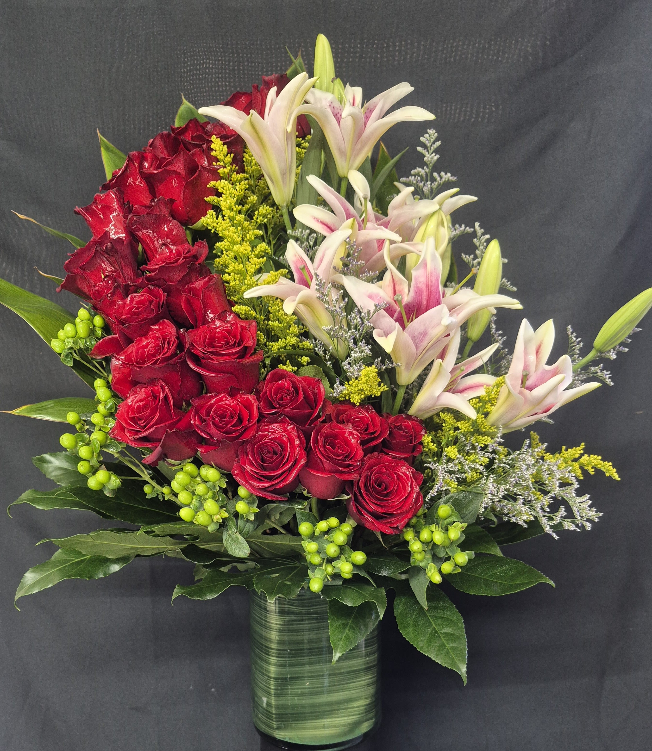 ROYAL RUBY RESERVE - A STRIKING 30 INCHES TALL ARRANGEMENT OF 24 STEMMS OF RICH RED ROSES AND SOFT PINK LILLIES, ACCENTED WITH VIBRANT GREEN HYPERICUM BERRIES AND FRESH GREENERY. DESIGNED IN A MODERN LEAF LINED GLASS CYLINDER VASE. THIS BOUQUET OFFERS A BEAUTIFUL BALANCE OF BOLD COLOR AND ELEGANT TEXTURE; PERFECT FOR ROMANTIC OCCASIONS OF MEANINGFUL CELEBRATIONS.