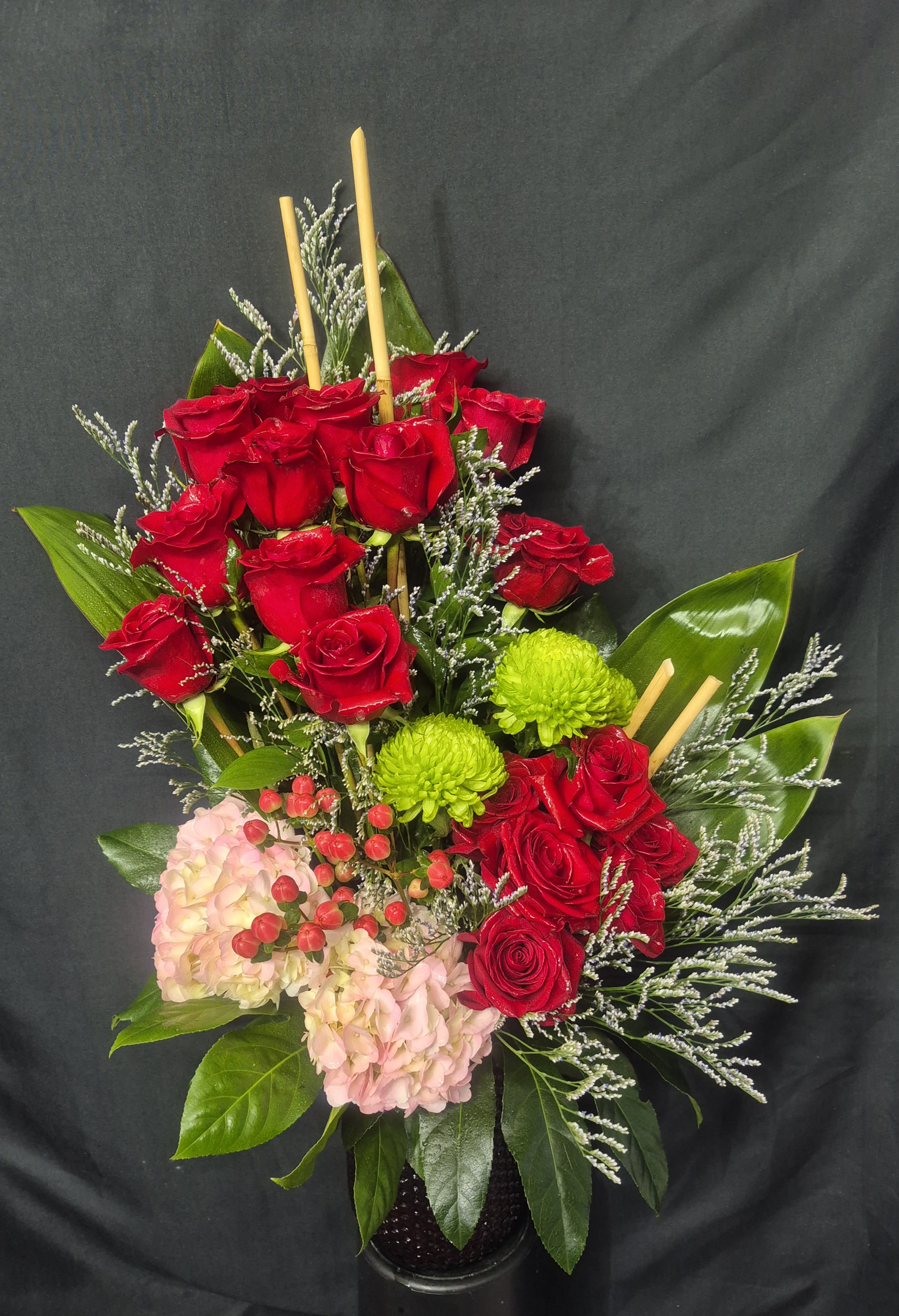 ROSE GARDEN DELIGHT - A striking and tall modern arrangement featuring vibrant red roses paired with soft pink hydrandes, green button mums, and textured accents. Designed with natural greenery and bamboo elements, it offers a bold but elegant presentation perfect for special occasions or heartfelt expressions.